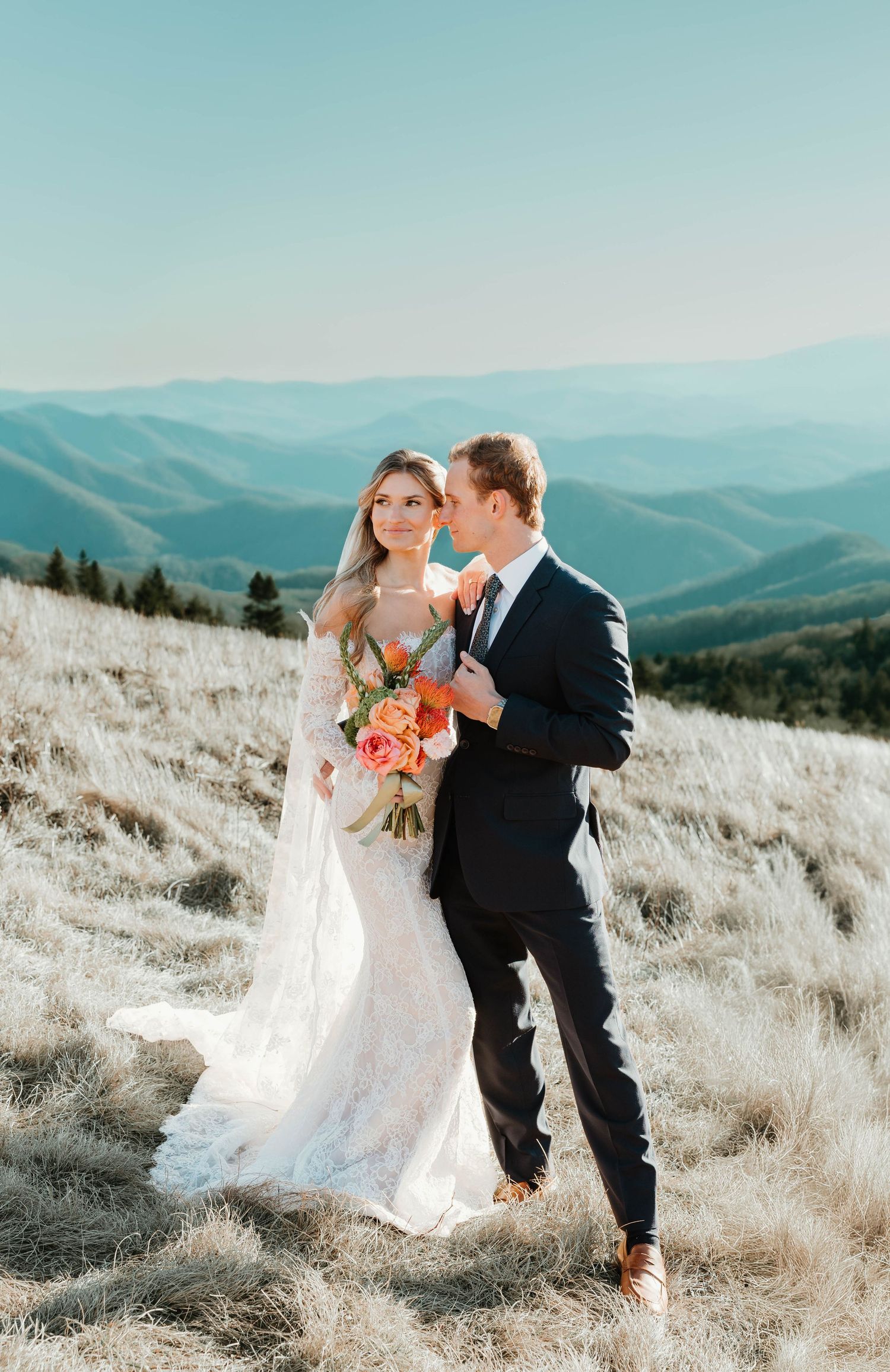 Newlywed couple shares romantic moments on a sunlit mountaintop with rolling hills and blue skies in the background.
