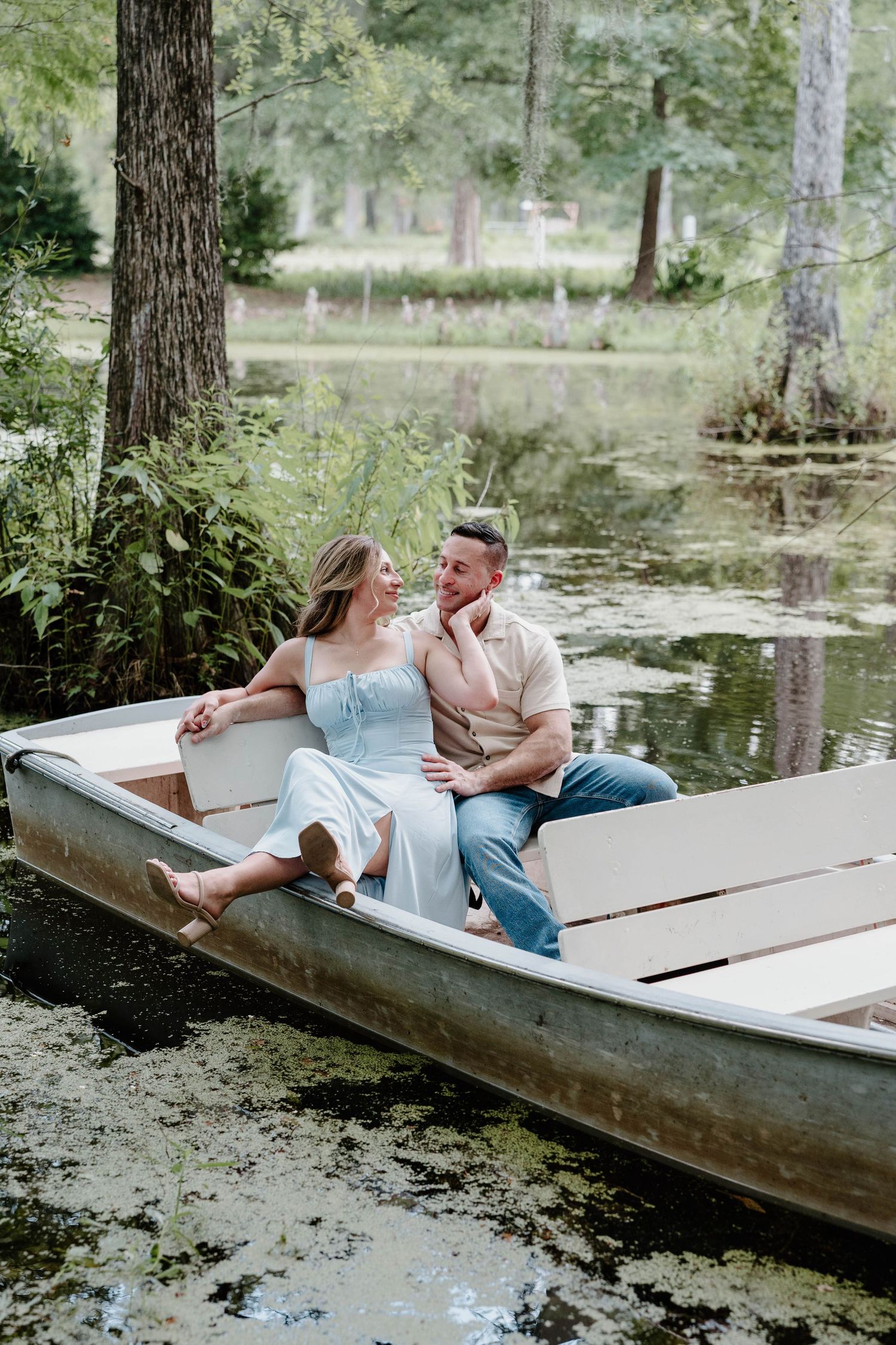 A couple relaxes together on a rustic wooden dock by a tranquil pond surrounded by trees on a summer day.