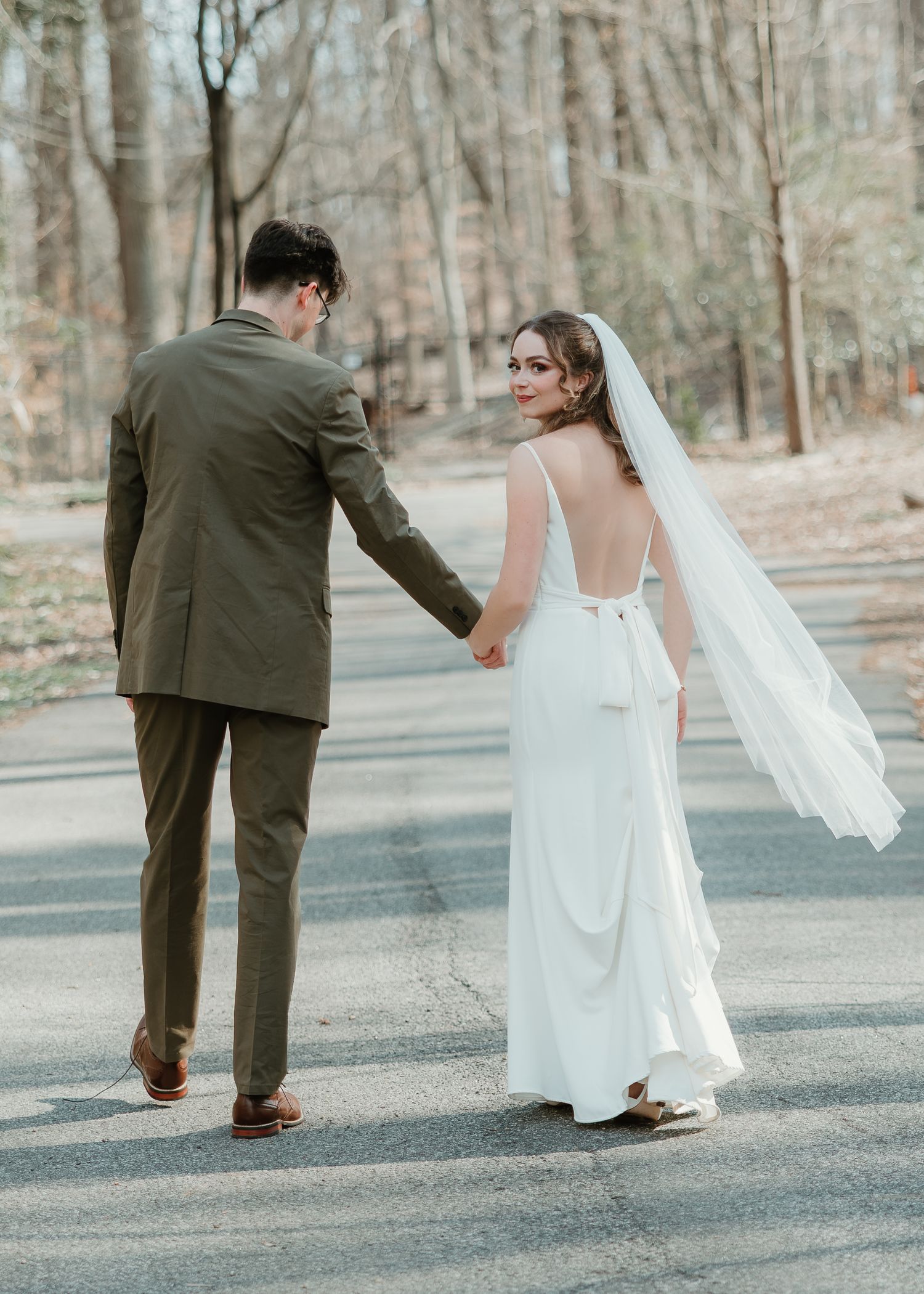 Bride in white gown and veil holds hands with groom in olive suit walking on a road surrounded by trees.
