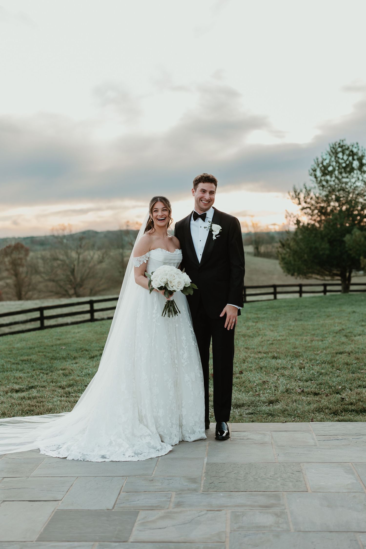 Bride in white lace gown holding bouquet stands with groom in black tuxedo on stone patio at sunset.