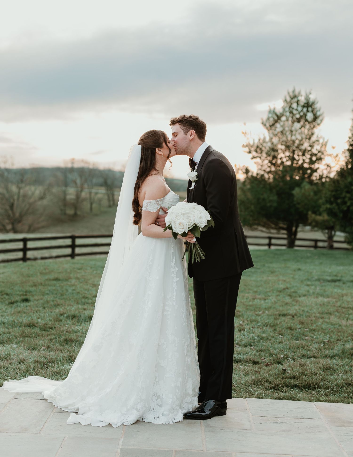 Bride and groom share a romantic kiss at sunset, with bride in lace gown holding white bouquet, groom in black tuxedo.
