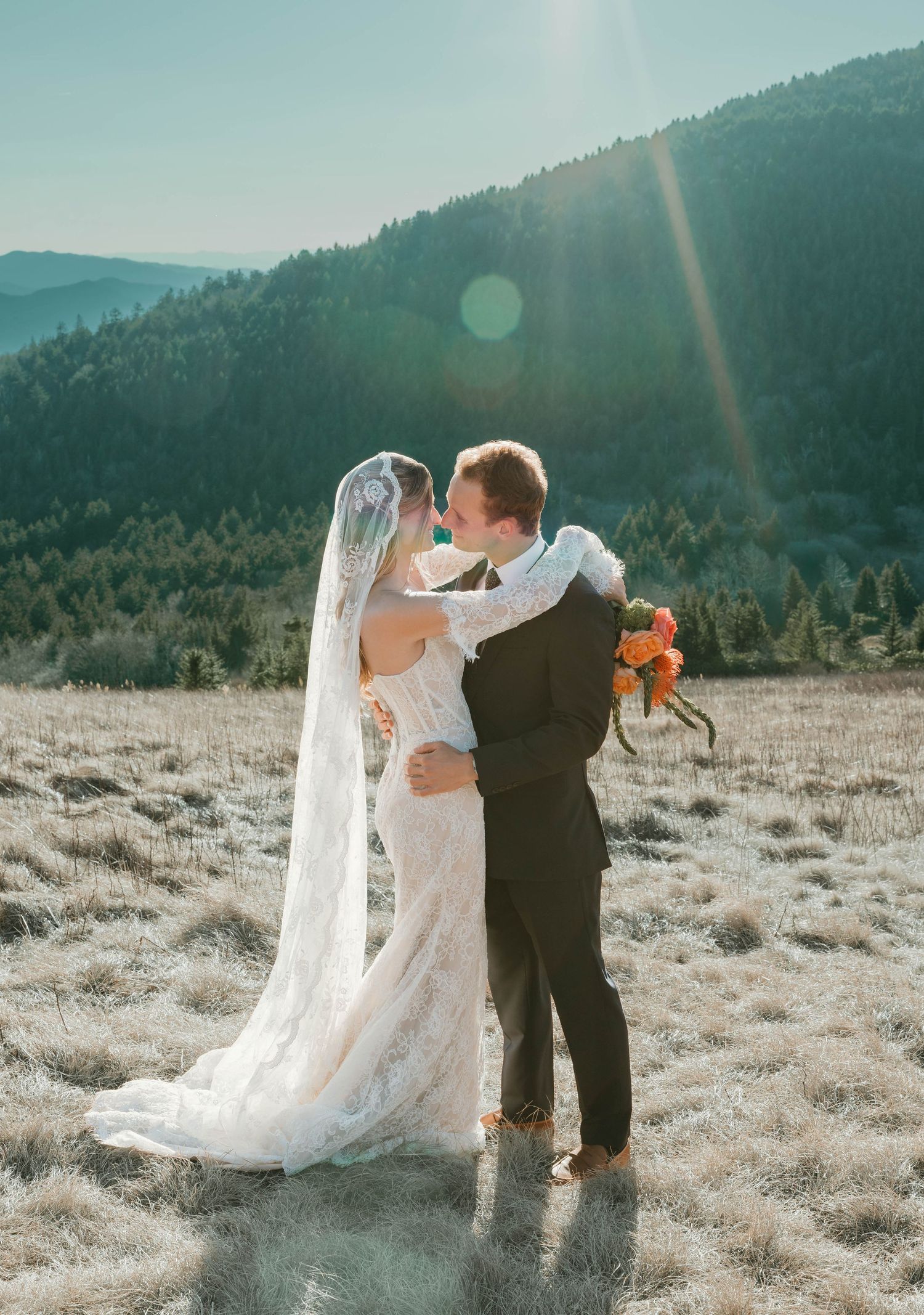 Romantic wedding couple embraces in a sunlit mountain meadow with tall grass and mountain vistas in the background.