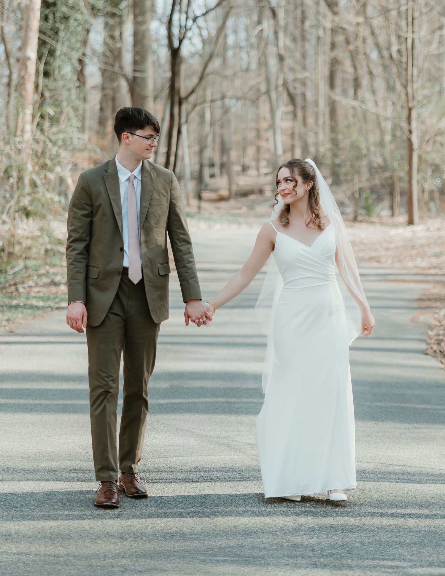 Bride and groom hold hands walking on a tree-lined path, she in a white gown with veil, he in an olive suit.