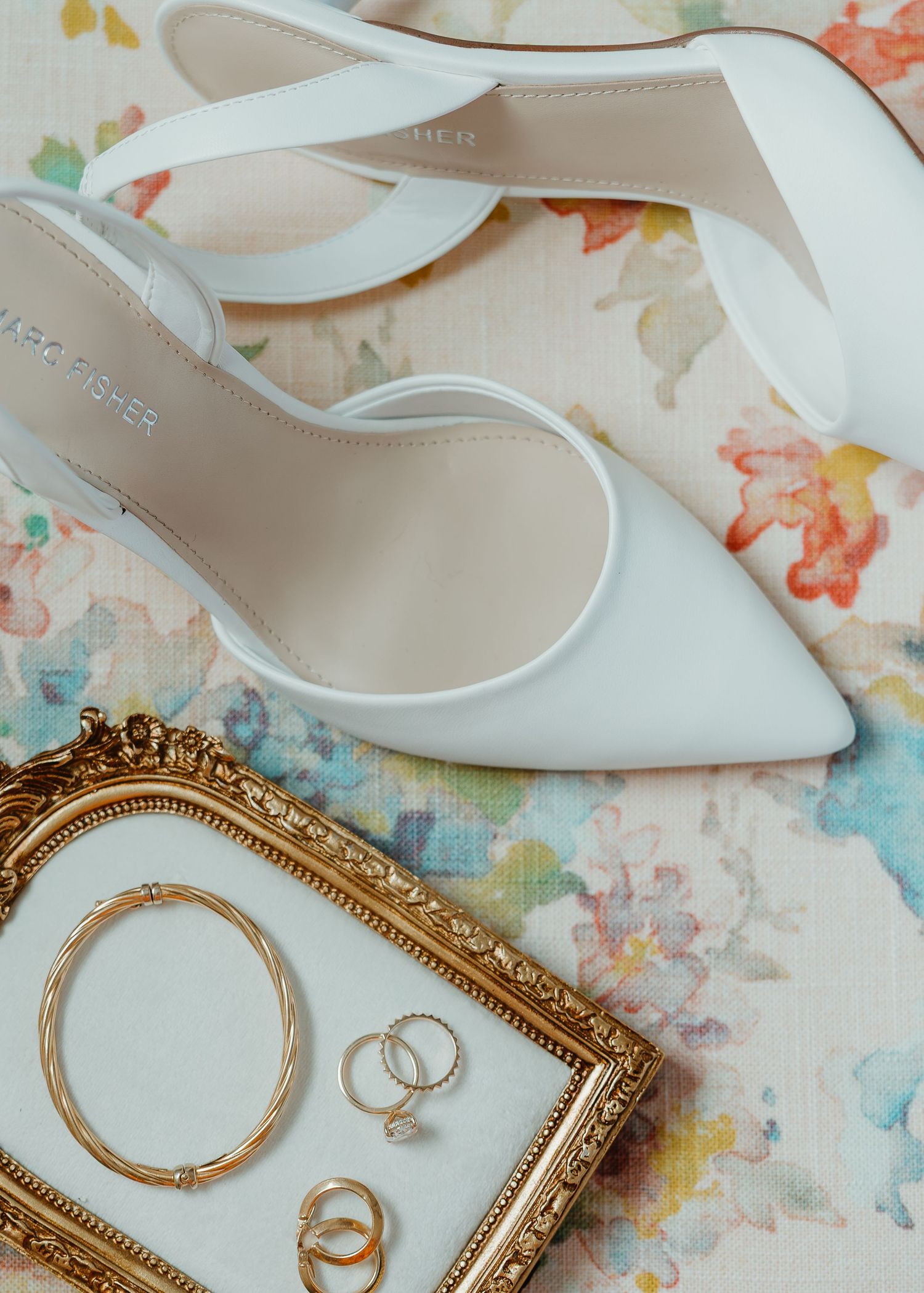 White Marc Fisher heels and gold jewelry including rings and bracelet displayed on floral fabric with ornate gold frame.