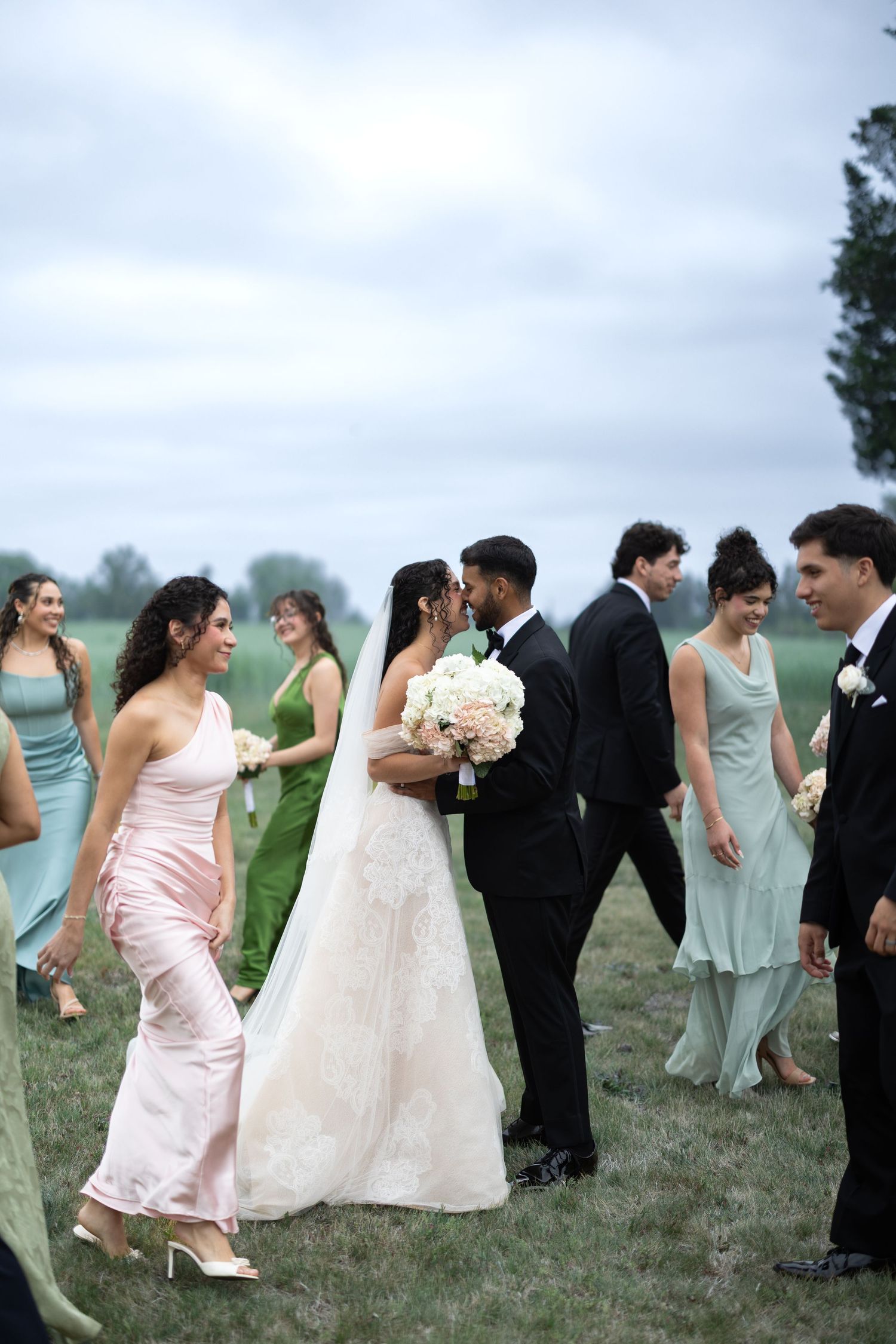 Bride and groom share a kiss surrounded by wedding party in mint and pink dresses on a green lawn.