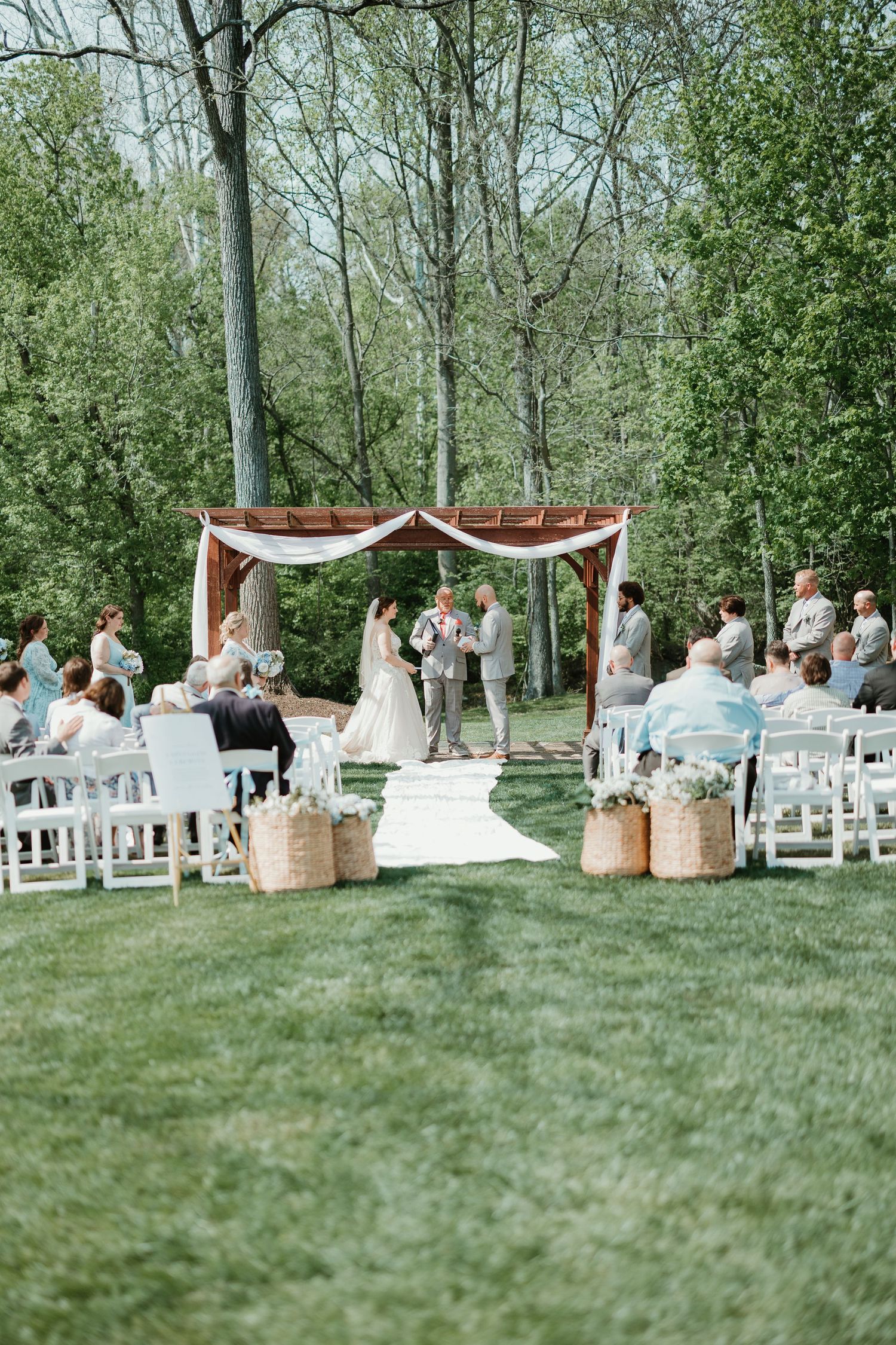 Outdoor wedding ceremony on green lawn with white chairs, floral arch, and couple exchanging vows surrounded by lush trees.