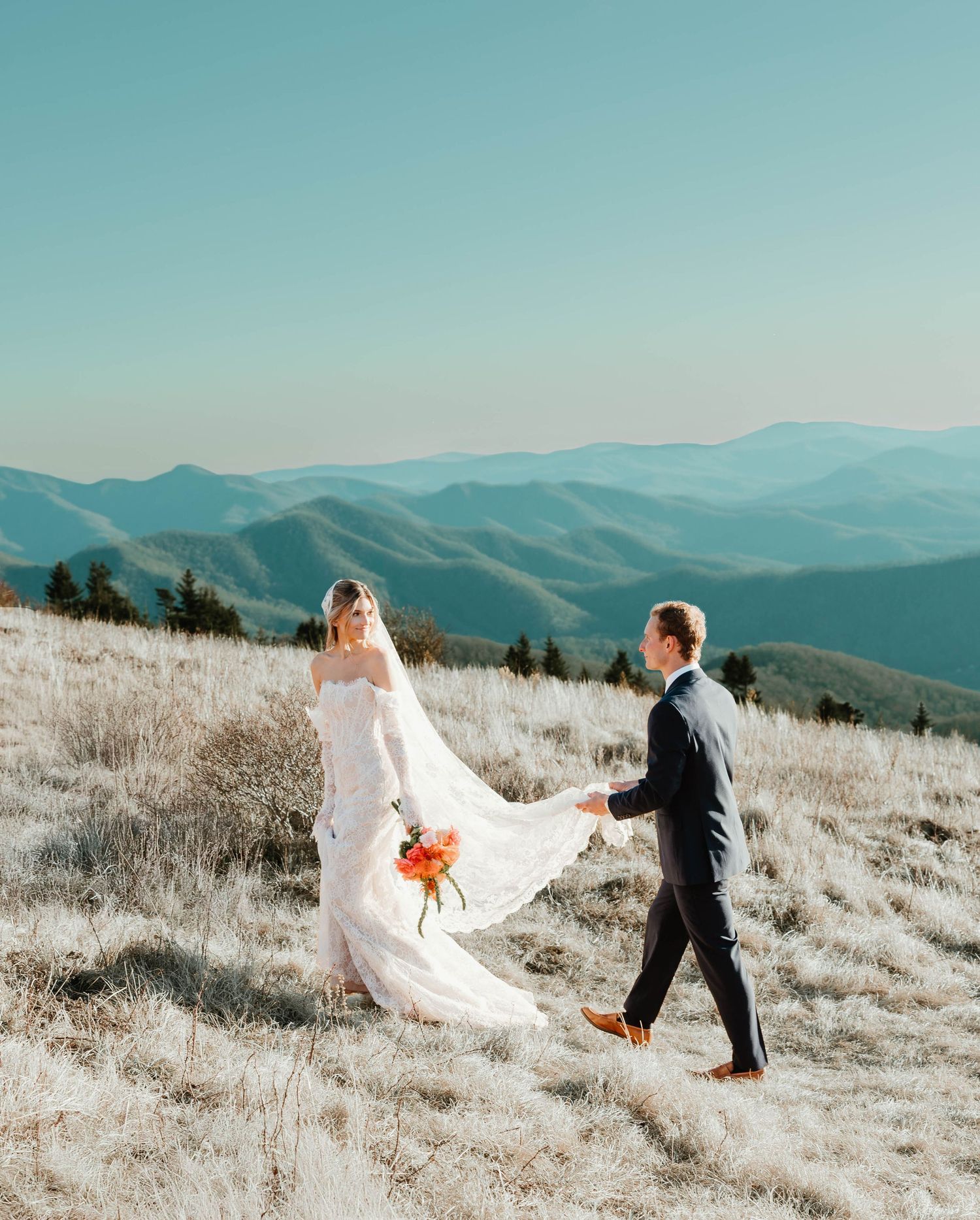 A couple shares a playful moment on a mountaintop meadow with rolling hills and mountains in the background.