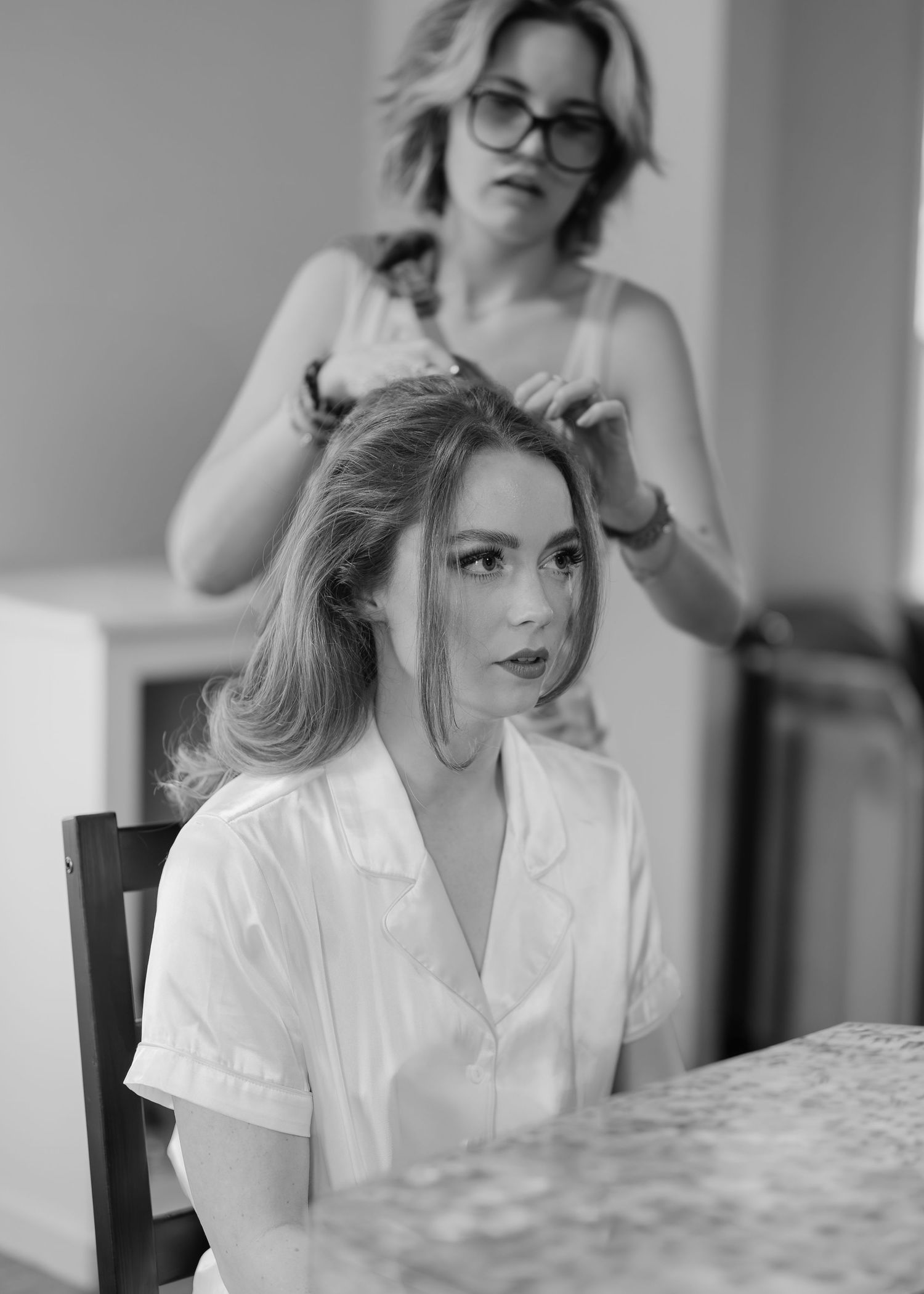 Black and white photo of a woman in white shirt getting her hair styled by a hairdresser wearing glasses.