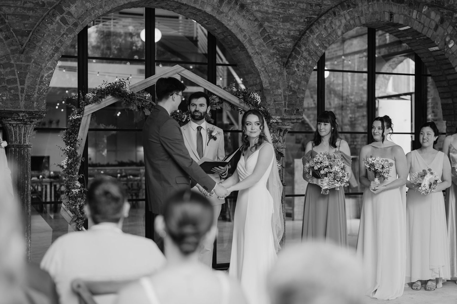 Black and white wedding ceremony photo with bride and groom exchanging vows, bridesmaids standing nearby in elegant venue.