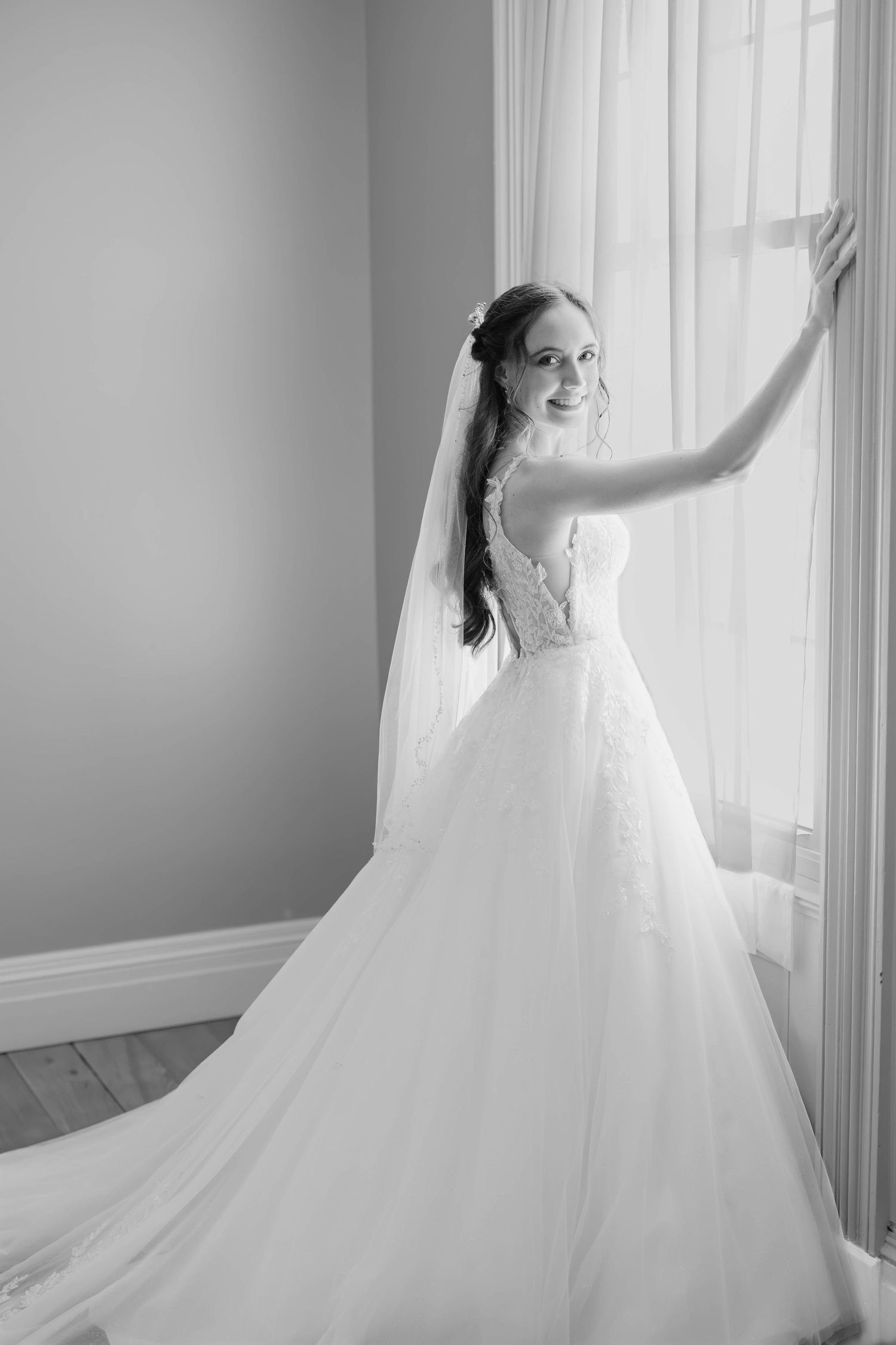 A bride in an elegant white tulle gown poses gracefully by a window in a black and white photograph.