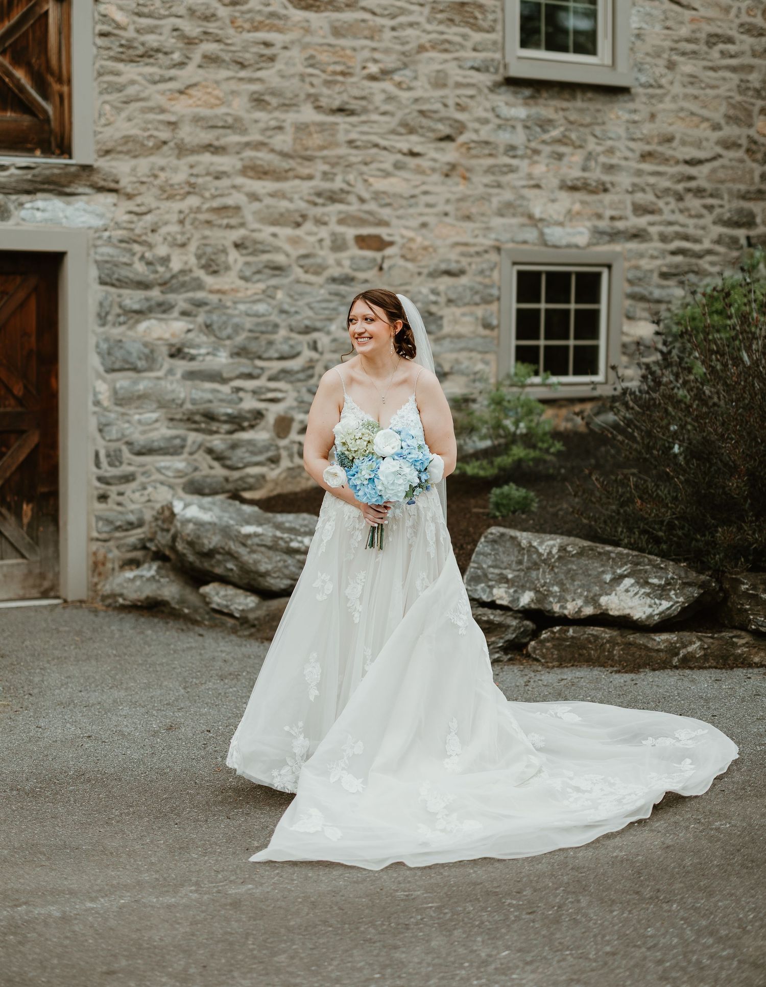 Bride in white gown with long train holds blue hydrangea bouquet outside rustic stone building.