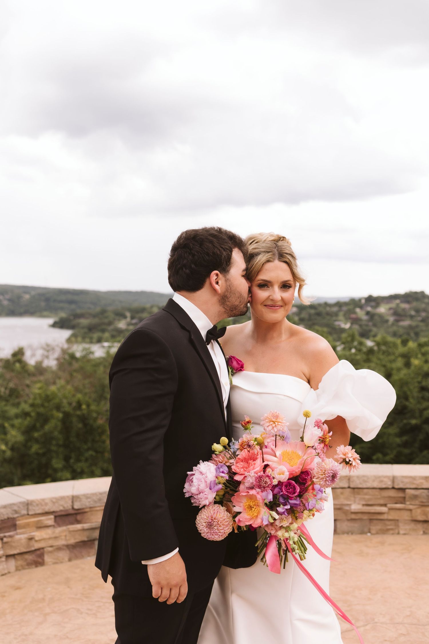 A wedding couple shares an intimate moment with pink and purple floral bouquet on a stone terrace.