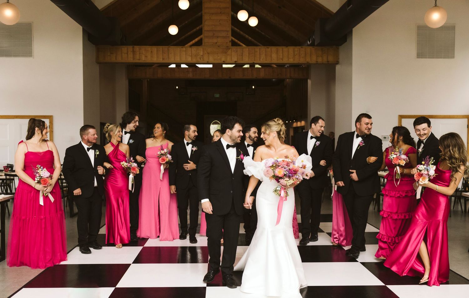A wedding party stands on a black and white checkered dance floor with bridesmaids in vibrant pink dresses.