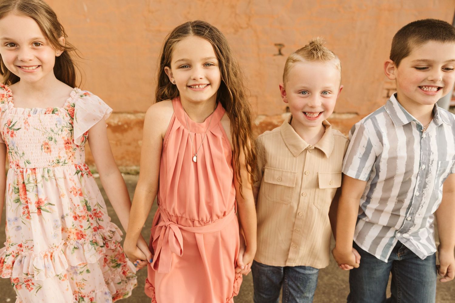 Group of children wearing pastel and floral outfits lined up against a tan wall.