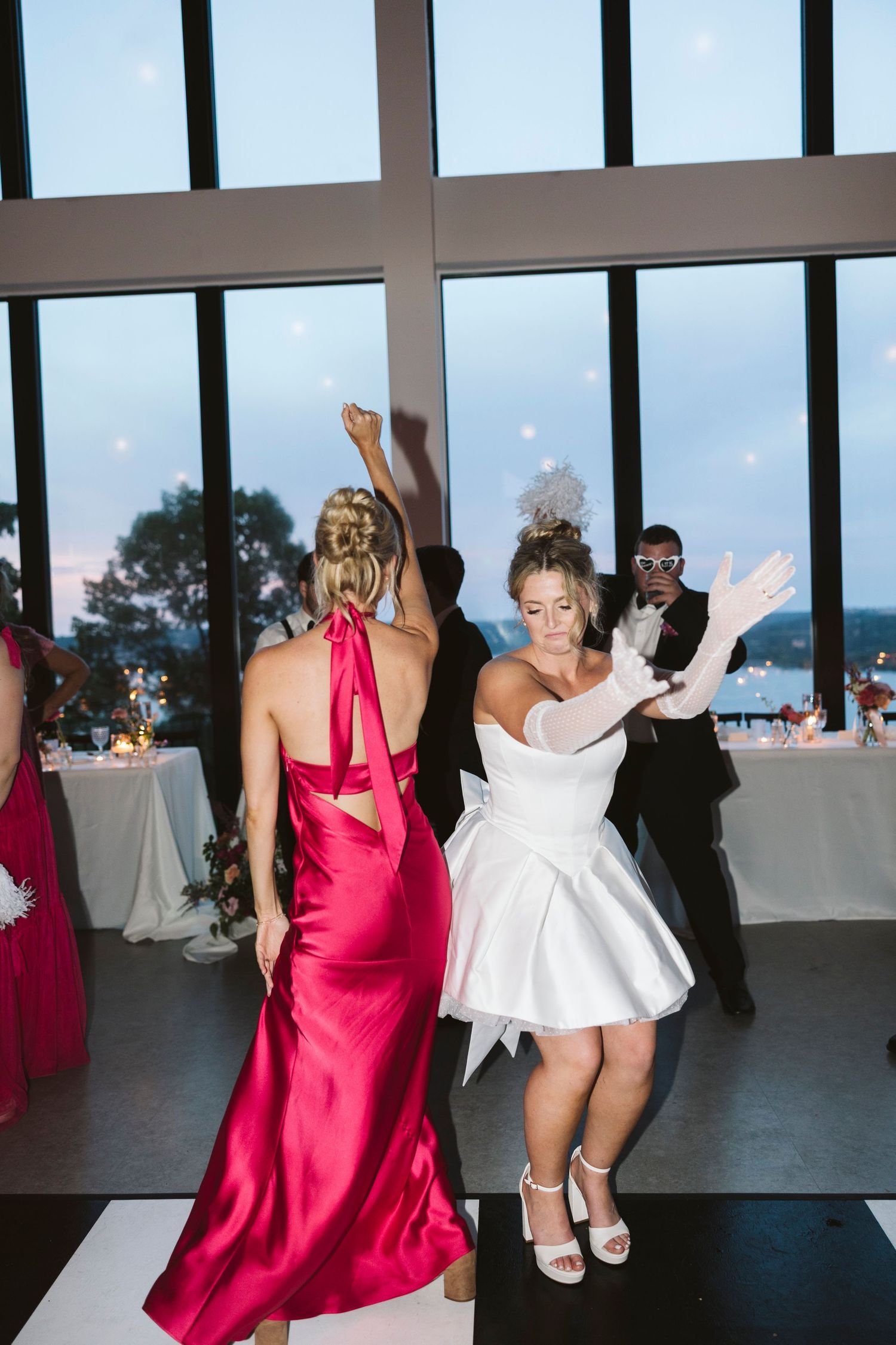 Wedding party members in pink dresses and white gowns dance energetically at an indoor reception.