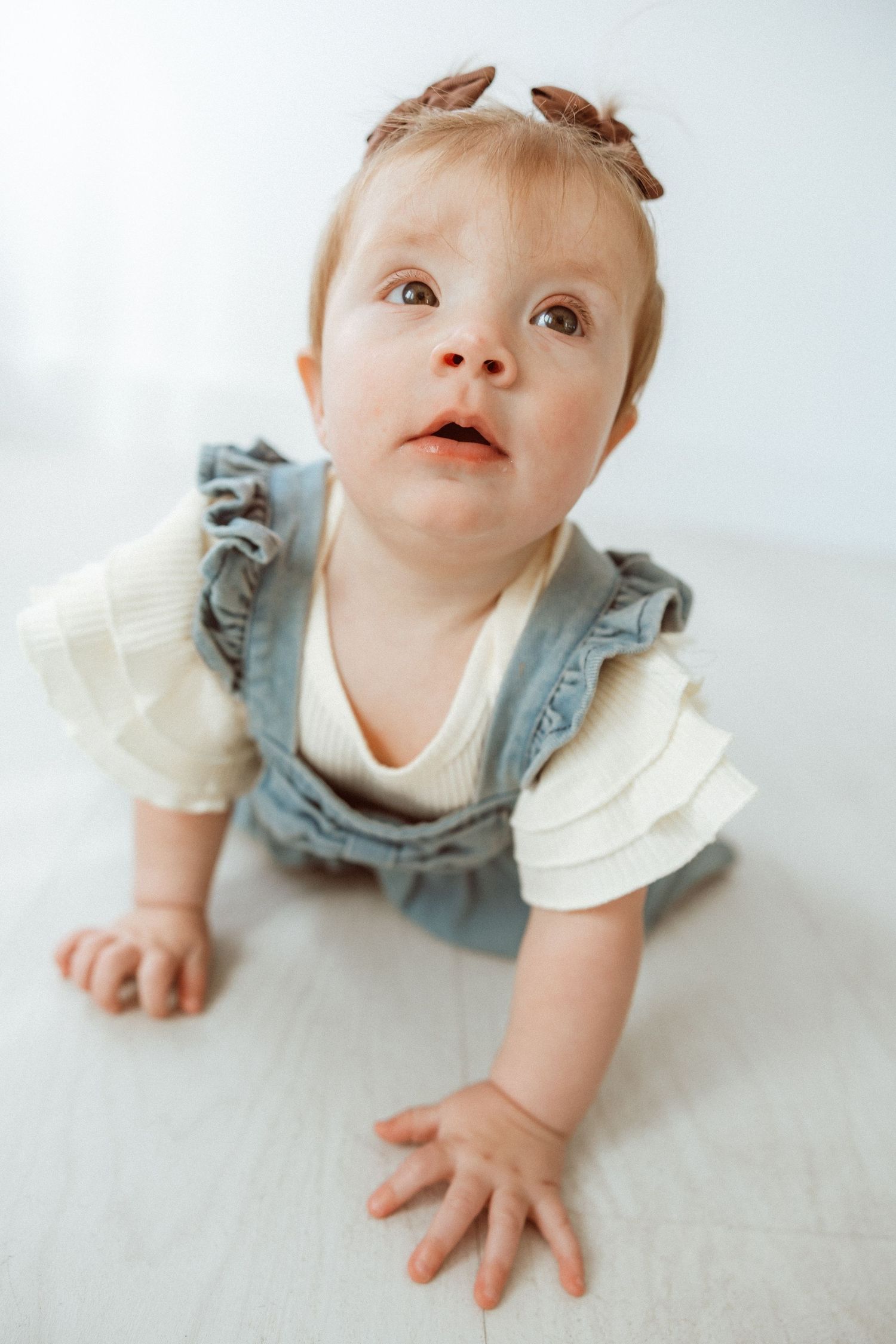 Baby in denim overalls and white top crawling on light floor.