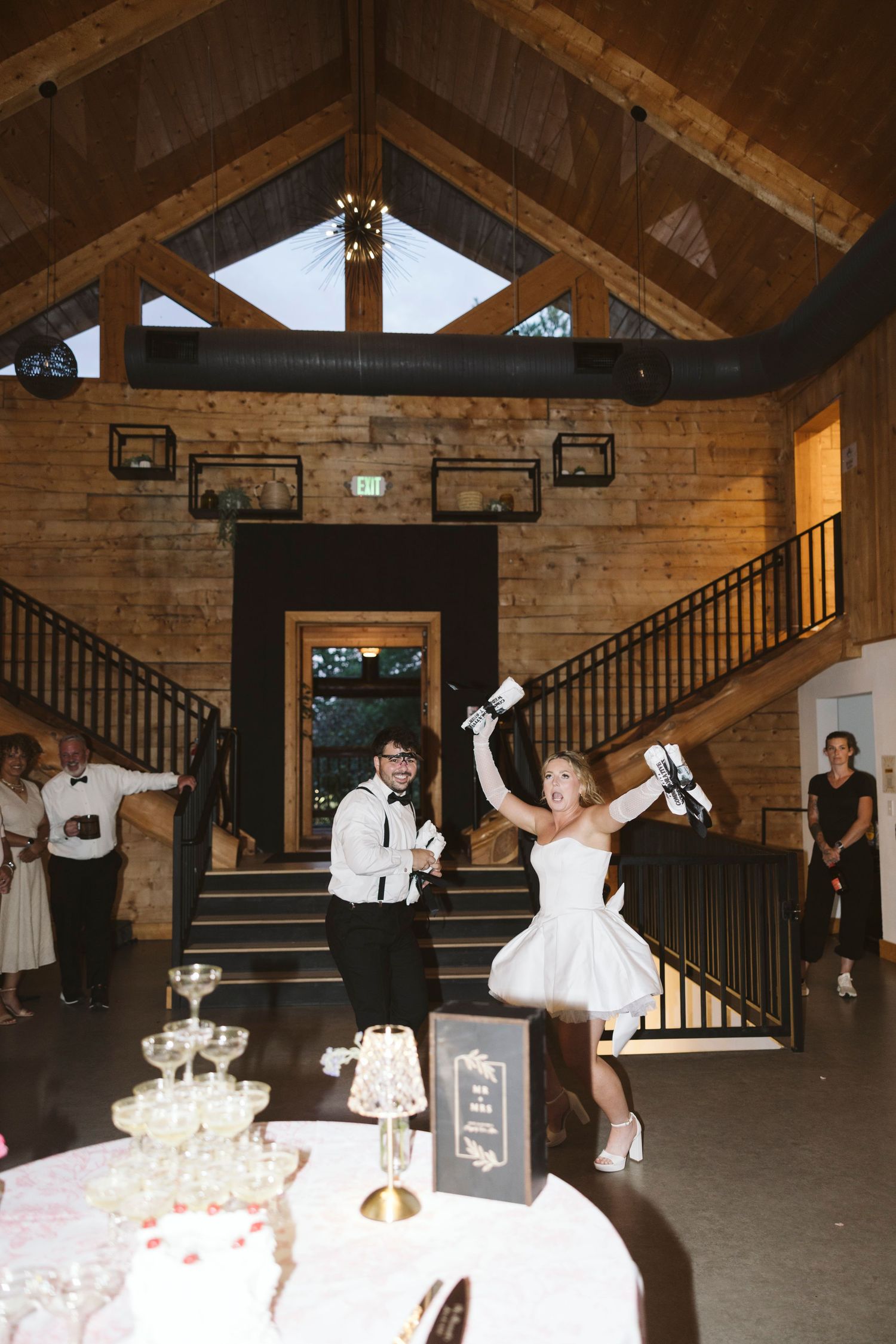Wedding party performs fun jumping action poses in a rustic barn venue with wooden beams and stairs.