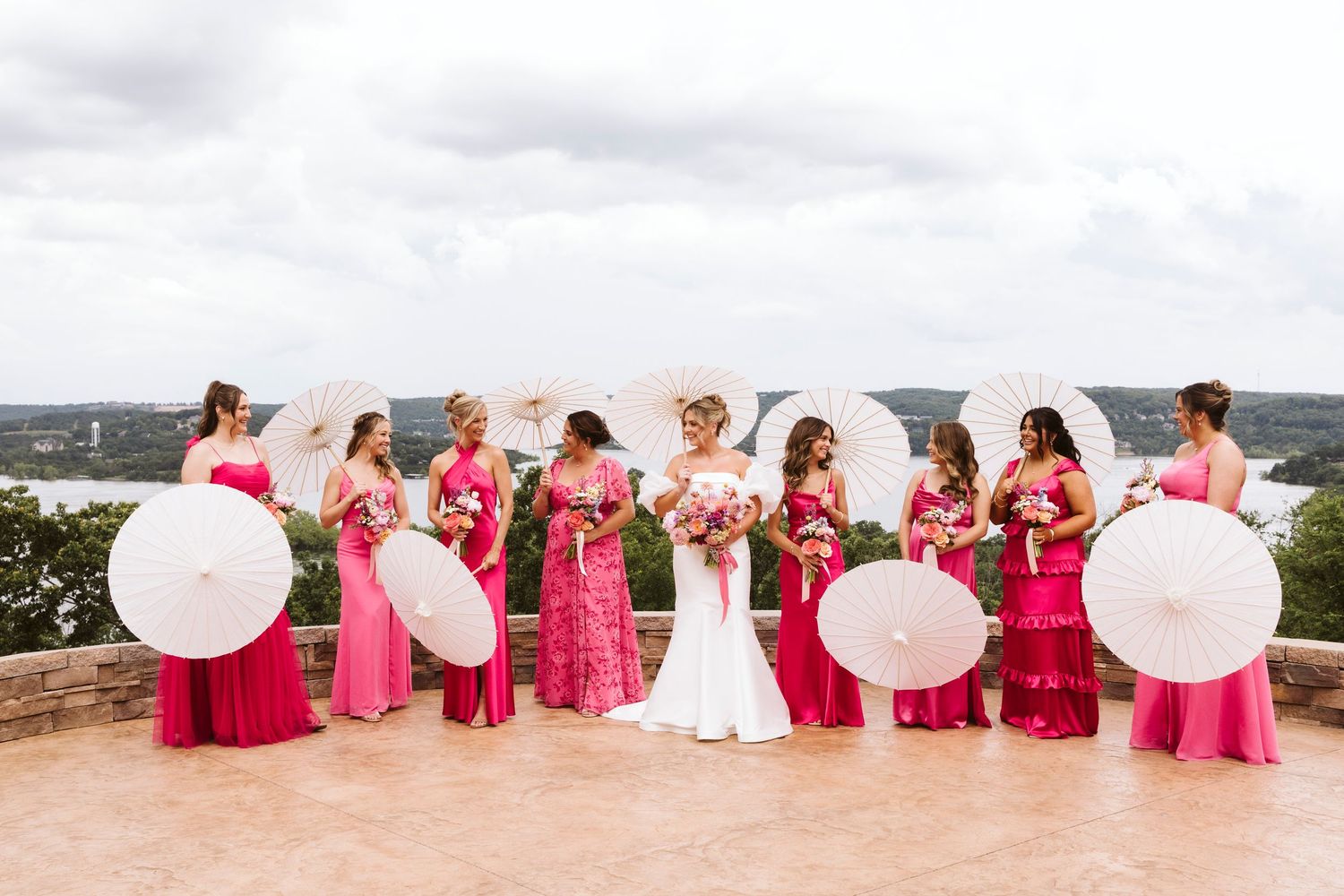 A bridal party in bright pink bridesmaid dresses holds white parasols while standing on a stone patio overlooking water.