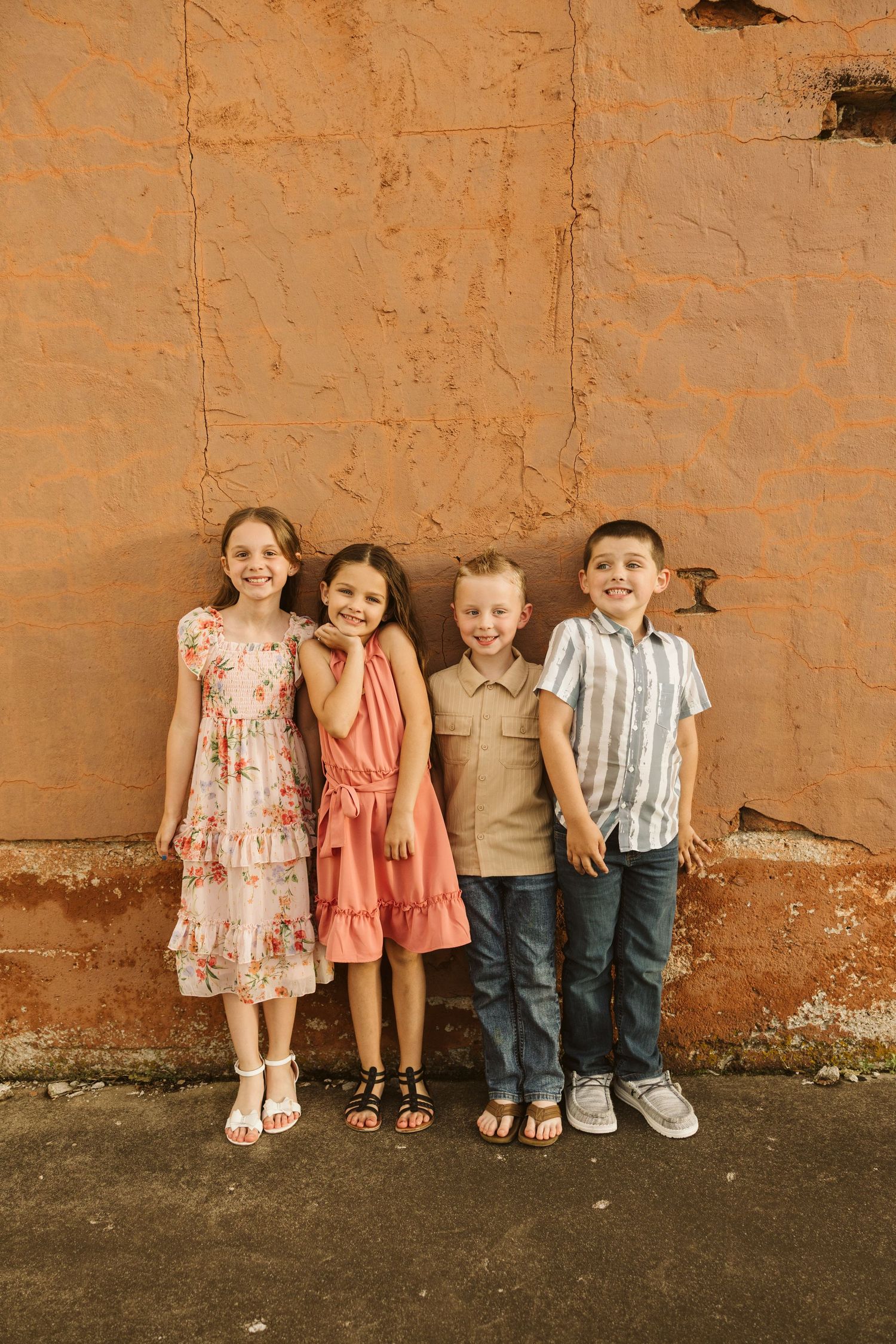 Four children stand against a textured orange wall wearing colorful summer outfits.