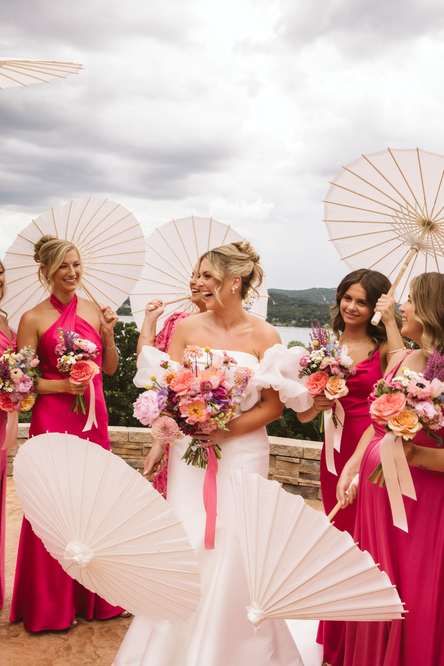Bridesmaids in bright pink dresses holding white parasols posing for photos.