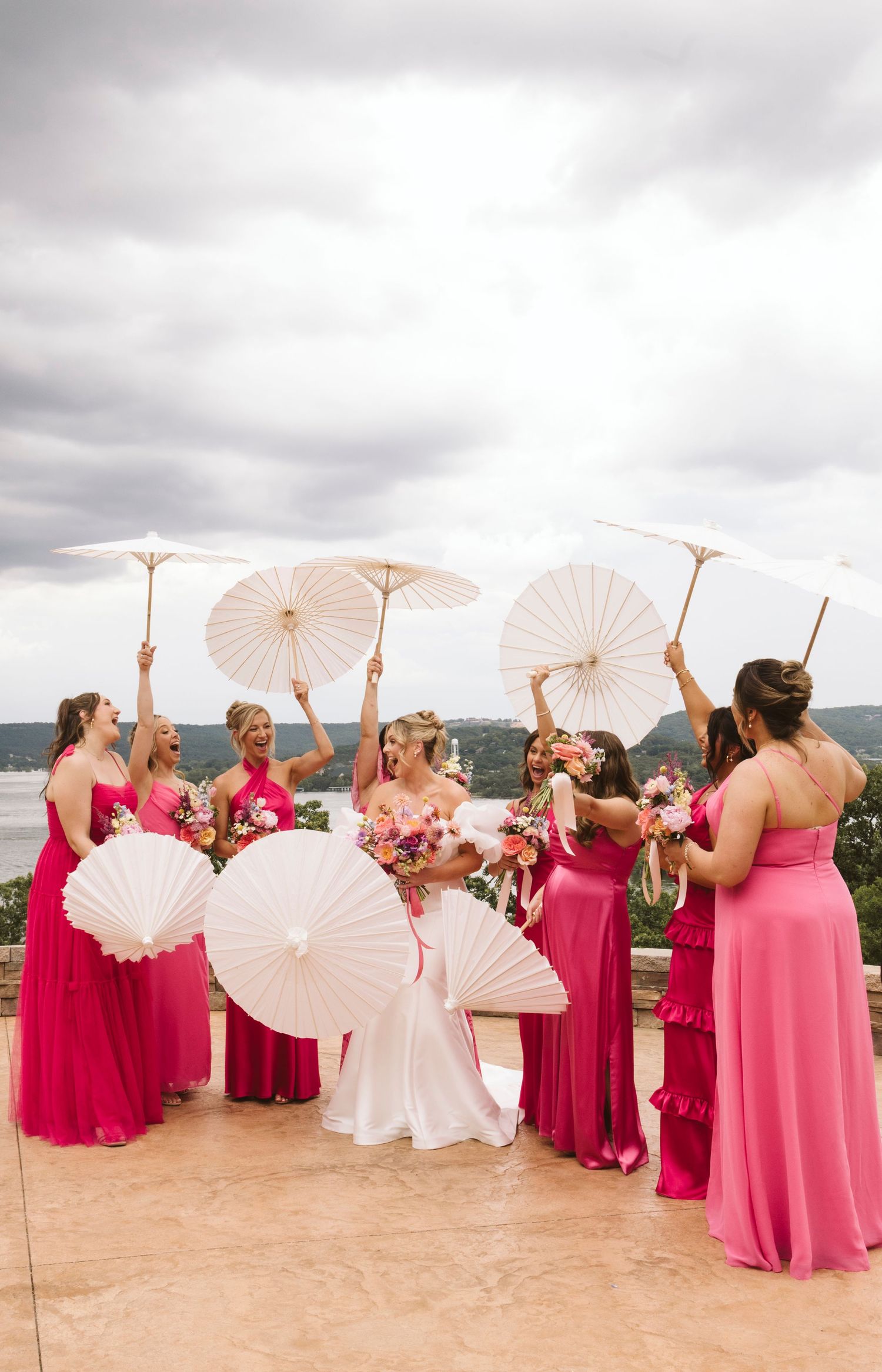 Bridal party in fuchsia dresses holding white parasols pose together on sandy outdoor setting.
