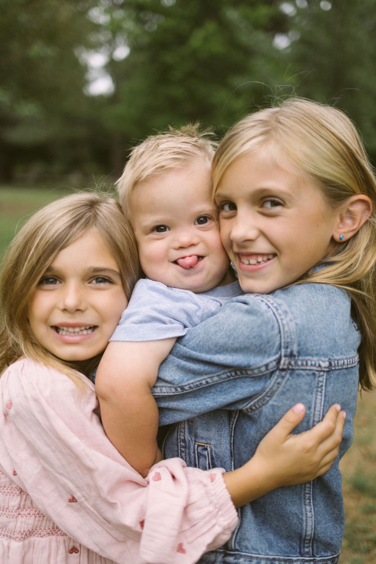 Three young children embrace and smile together outdoors in casual clothing.