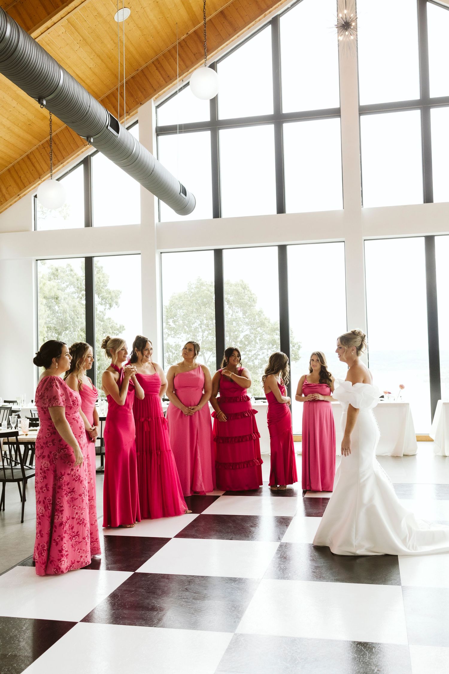 Bridesmaids in vibrant pink dresses gather in a modern venue with vaulted wooden ceiling.