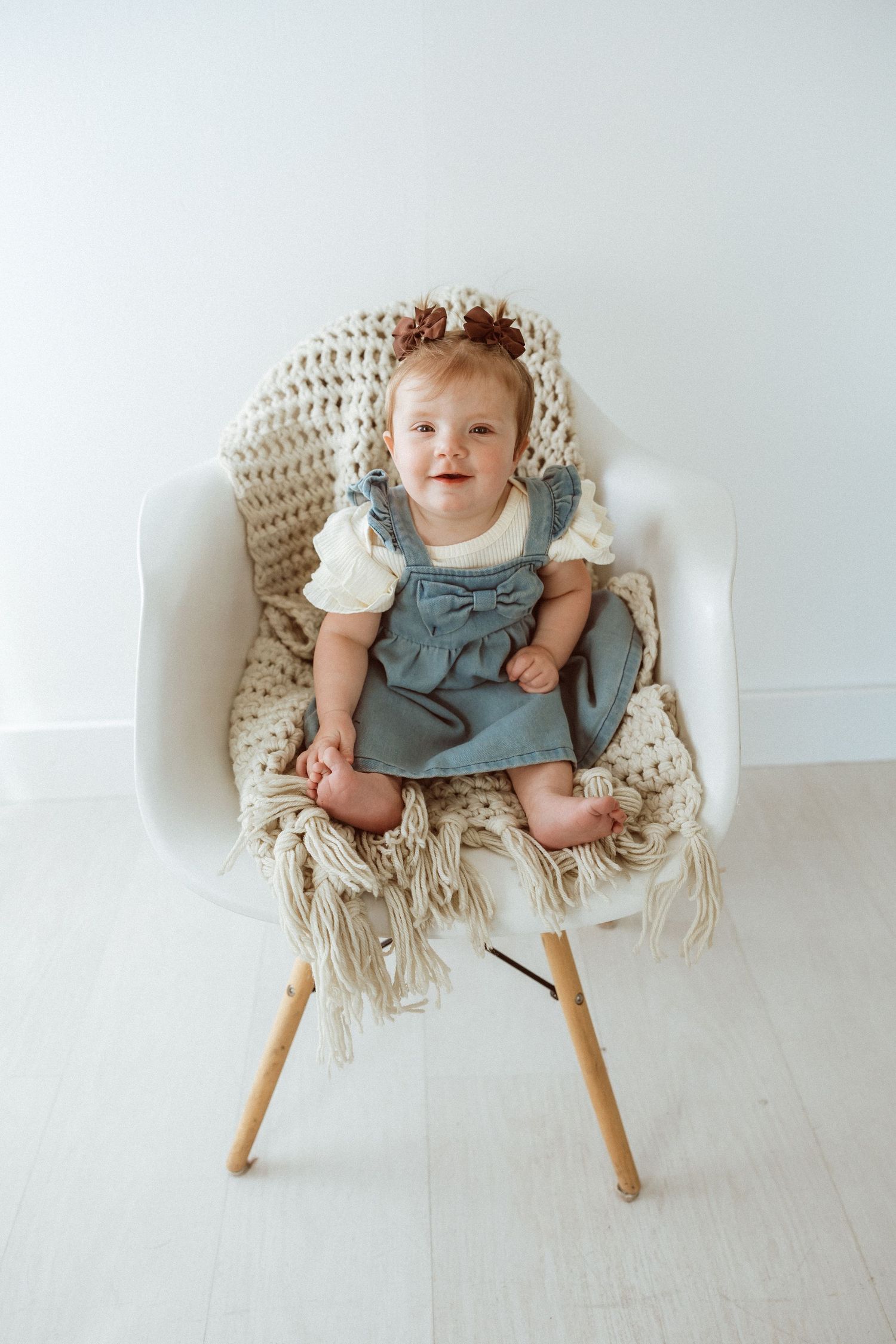 Baby sitting in white modern chair with knit blanket wearing blue dress.