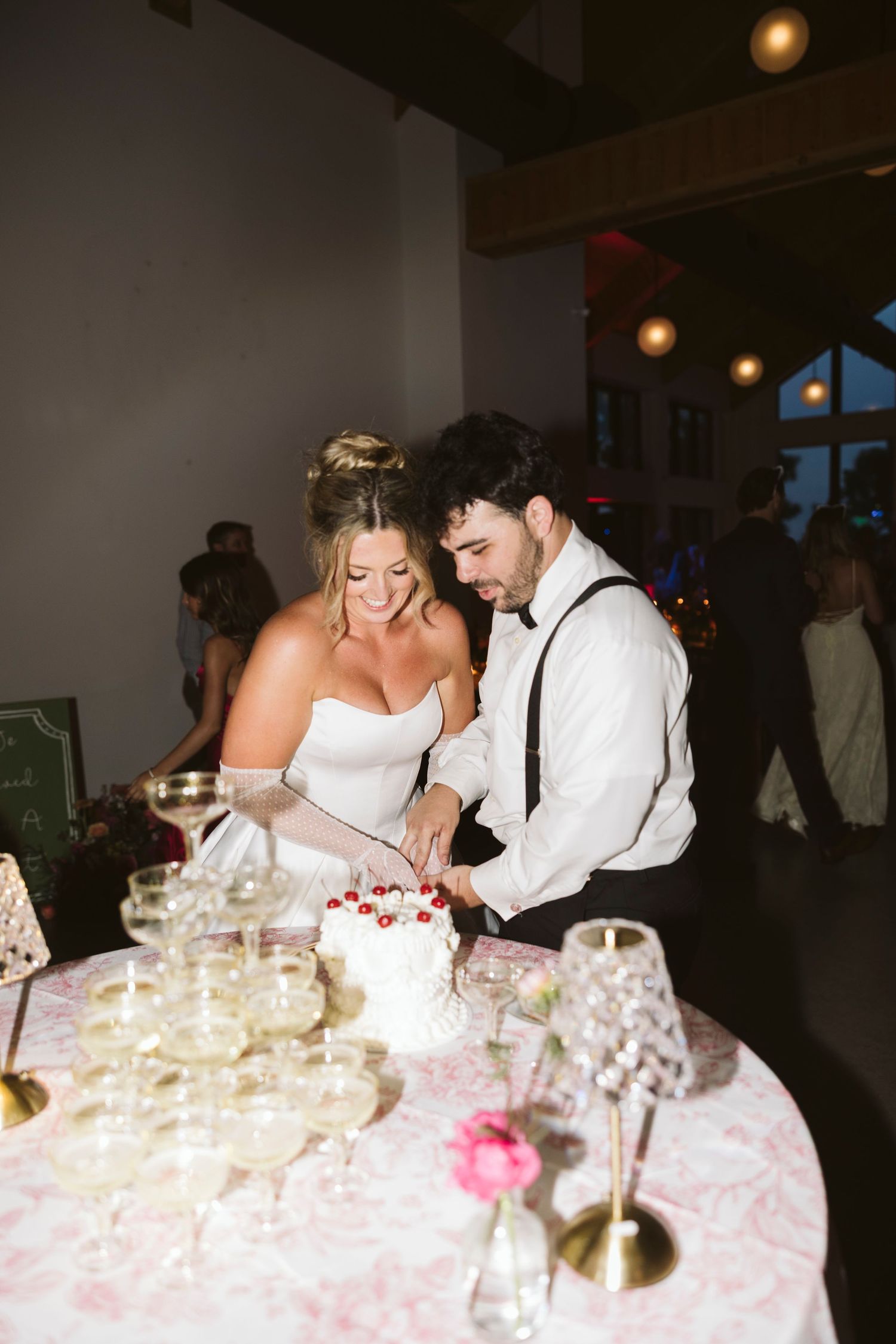 A couple shares a joyful moment while cutting their wedding cake at a reception with ambient lighting.