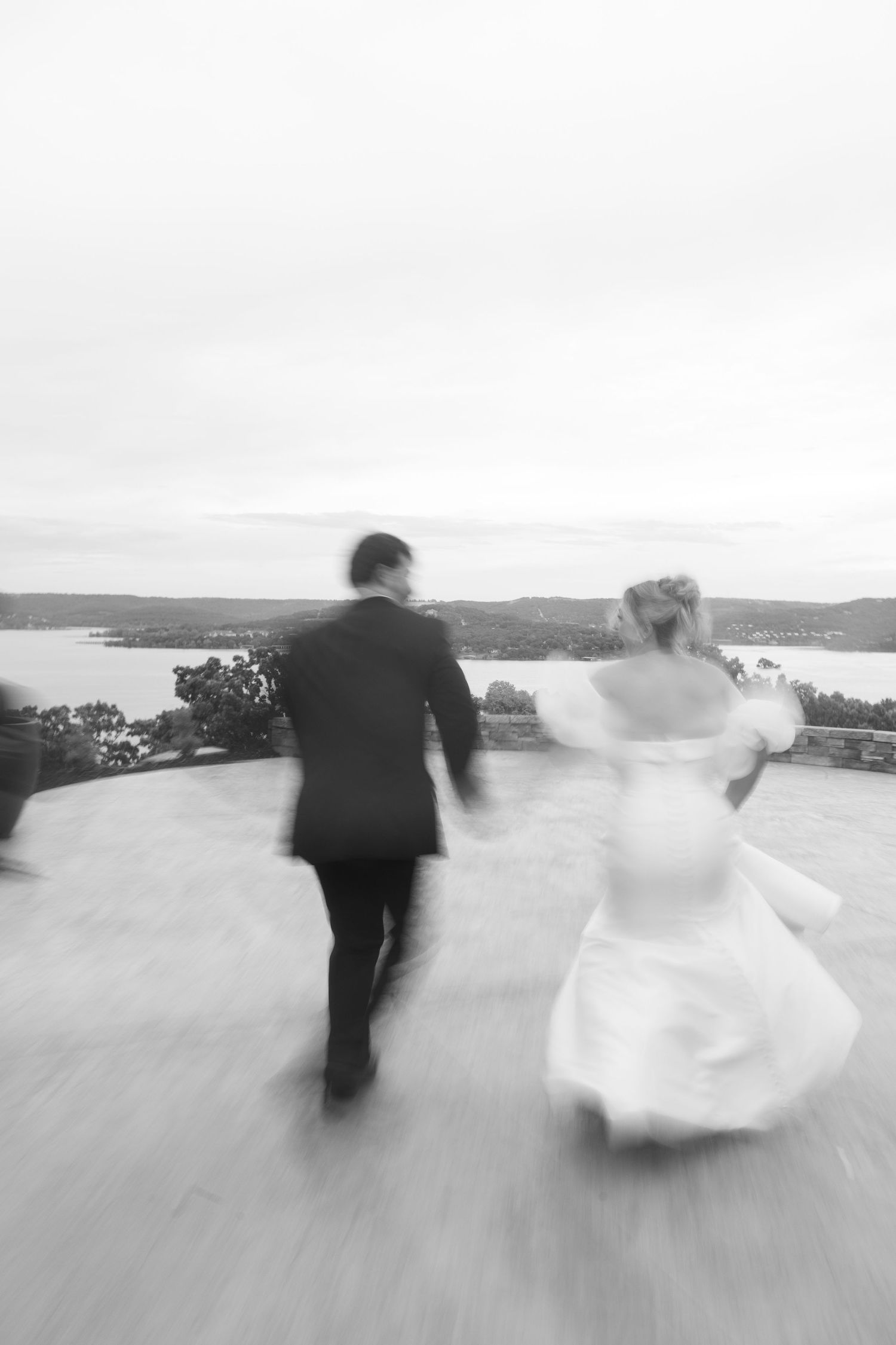 A black and white motion-blur photo of newlyweds running with a waterfront view.