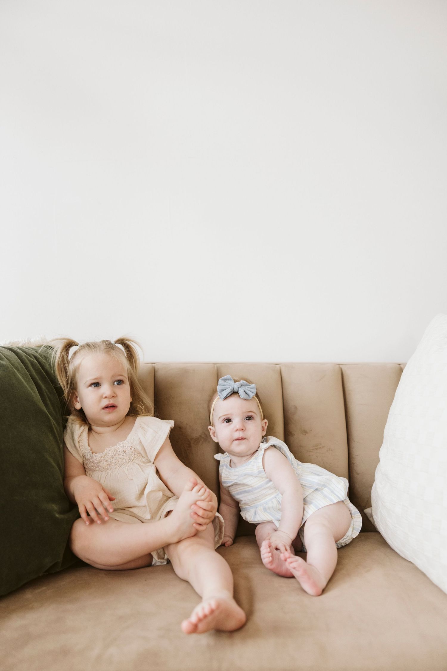 Three babies in light colored clothing sit together on a beige couch.
