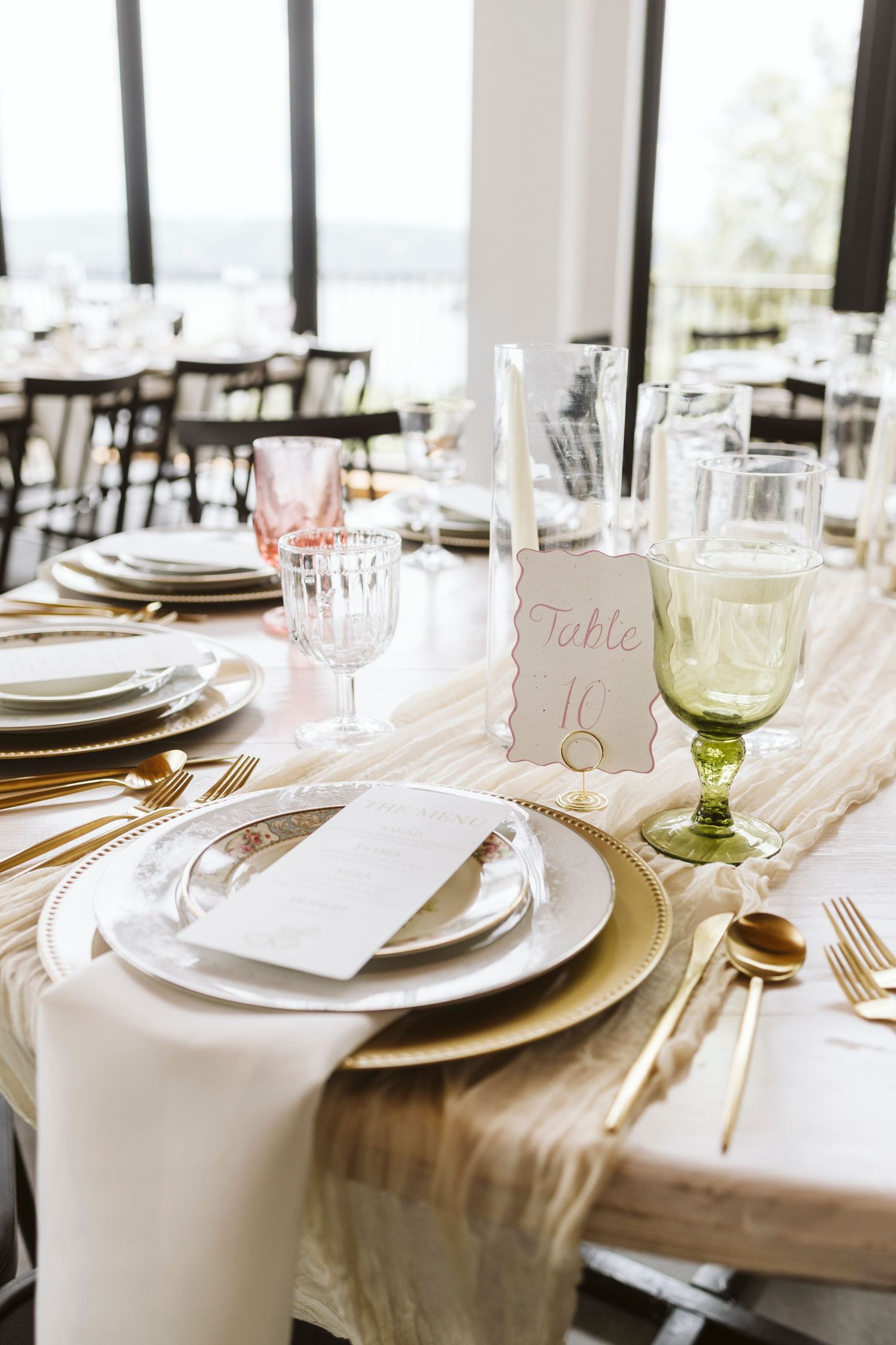 Elegant wedding table setting with white linens, gold charger plates, and soft green glassware on a rustic wooden table.