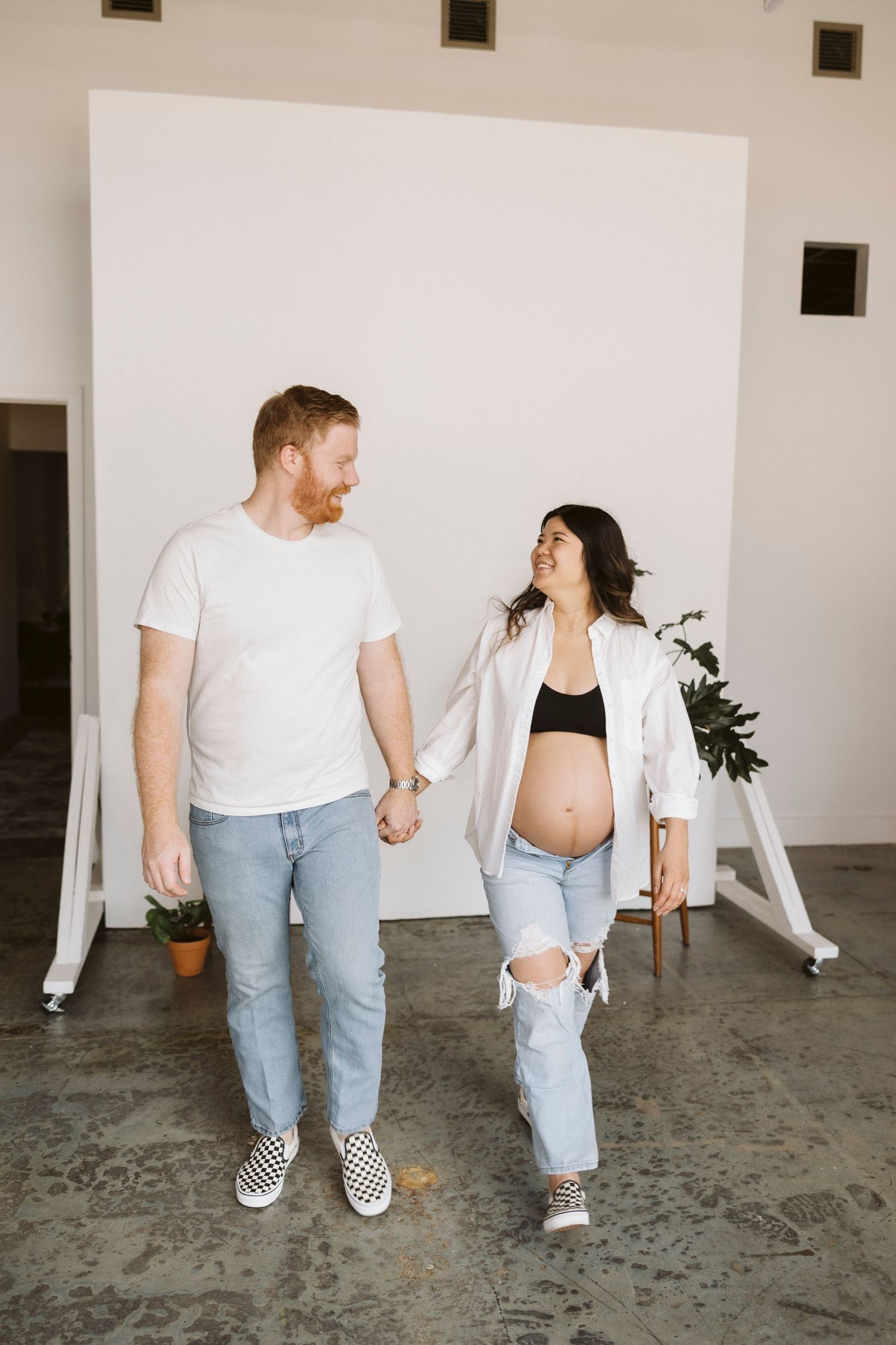 Expectant couple in casual wear holding hands against white backdrop during maternity photoshoot.