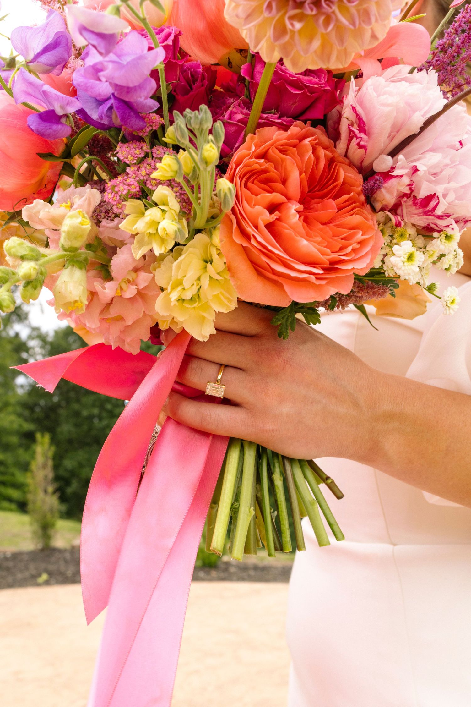 A vibrant wedding bouquet with coral roses, purple flowers and pink ribbons held by a hand with a wedding ring.