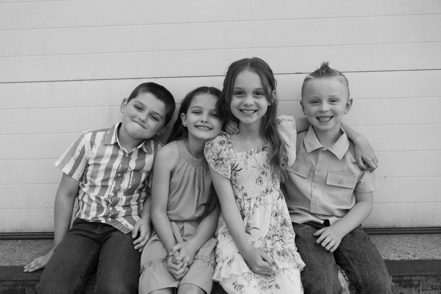 Black and white portrait of four siblings sitting casually together against a white wall.