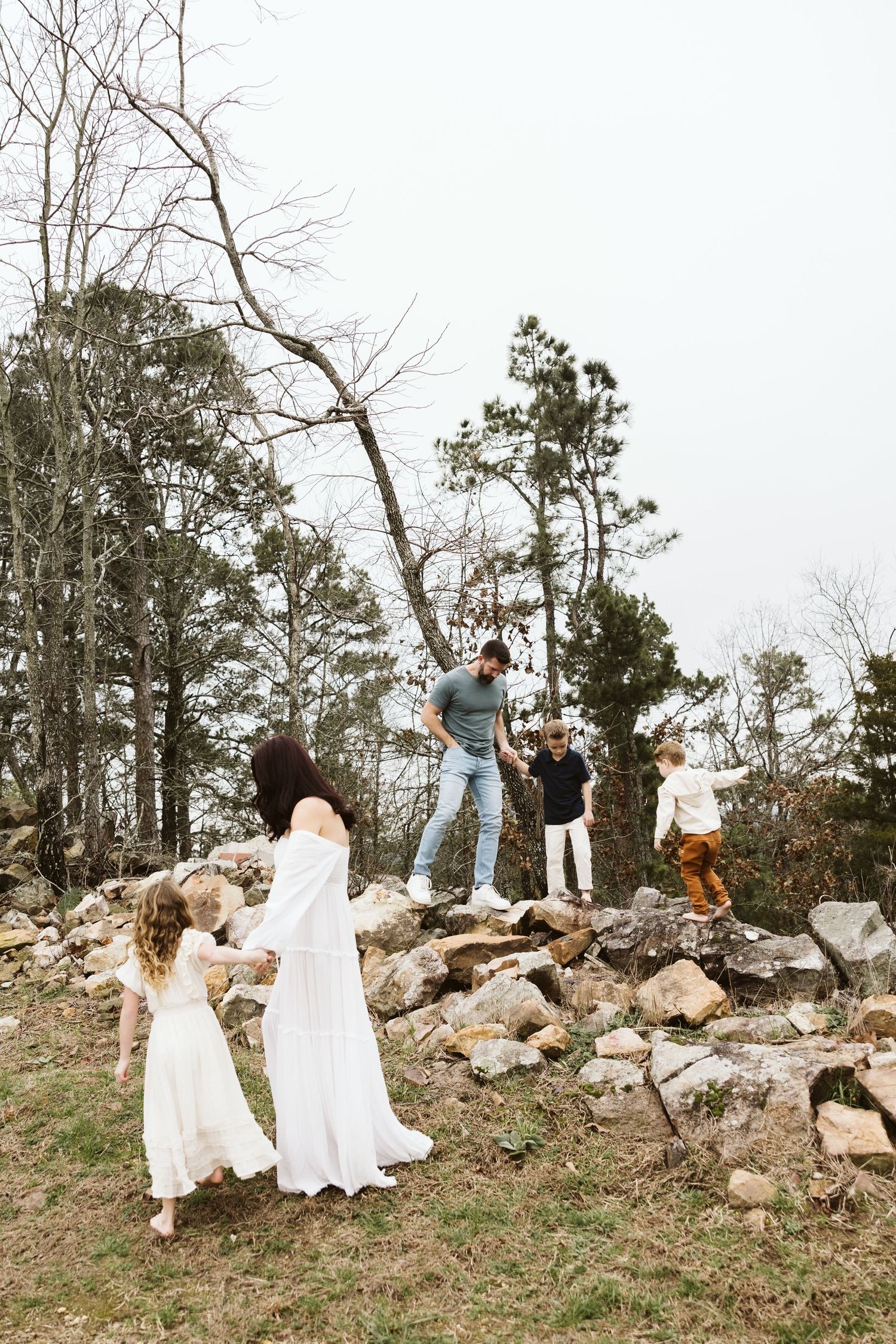 A group in flowy white dresses stands together on rocky terrain with tall trees in the background.