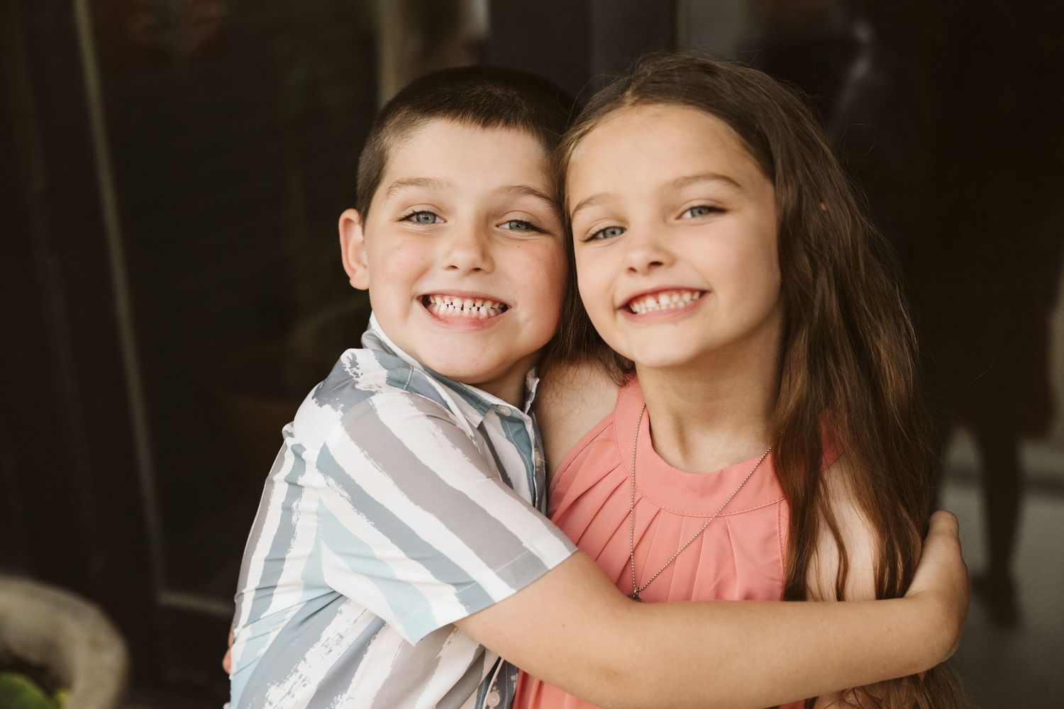 Two young children in white and coral colored outfits embrace and smile while posing for a heartwarming photo series.