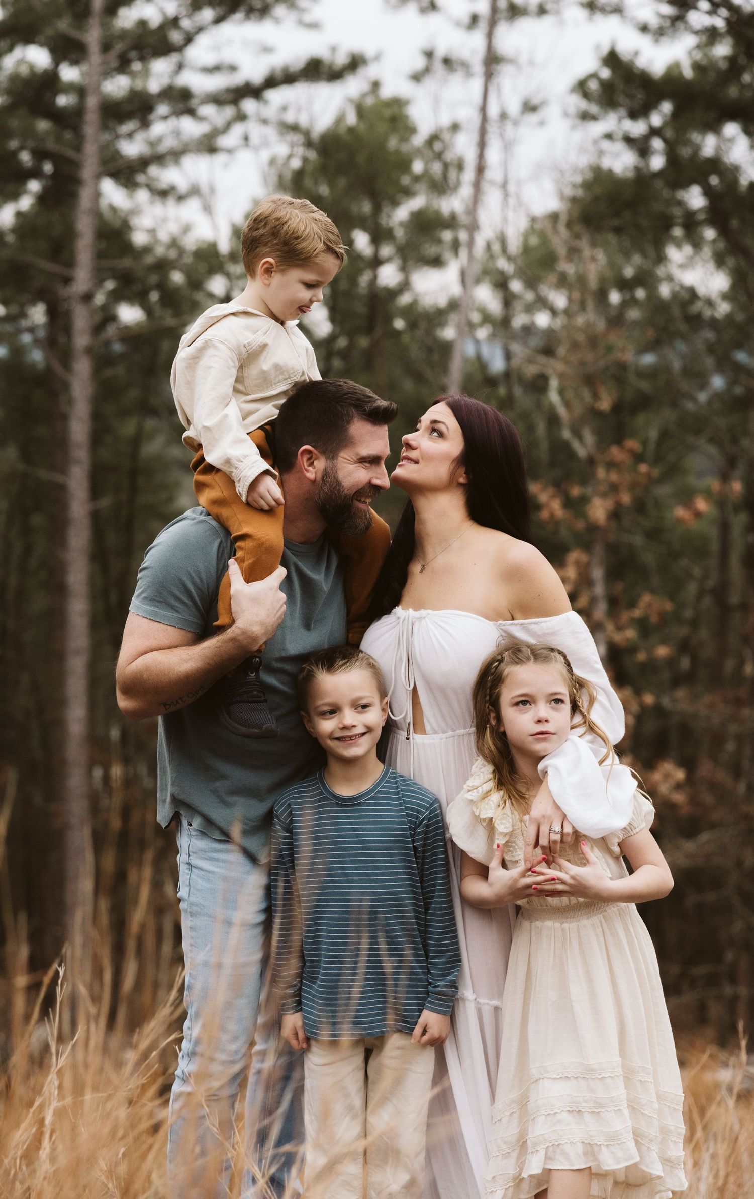 Outdoor family portrait in natural setting with pine trees and tall grass.