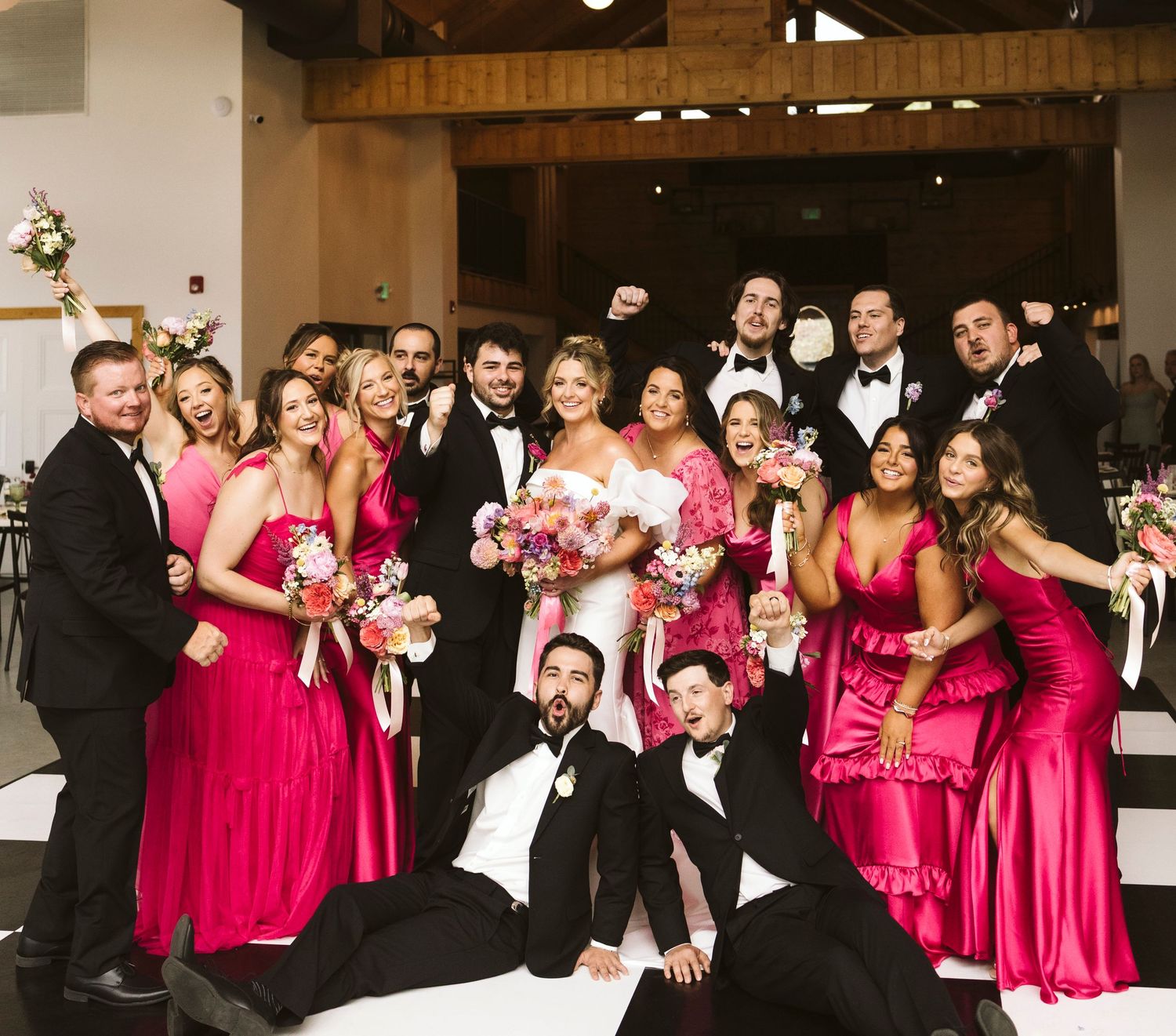 Wedding party in bright pink bridesmaid dresses and black tuxedos pose together for formal group photos.