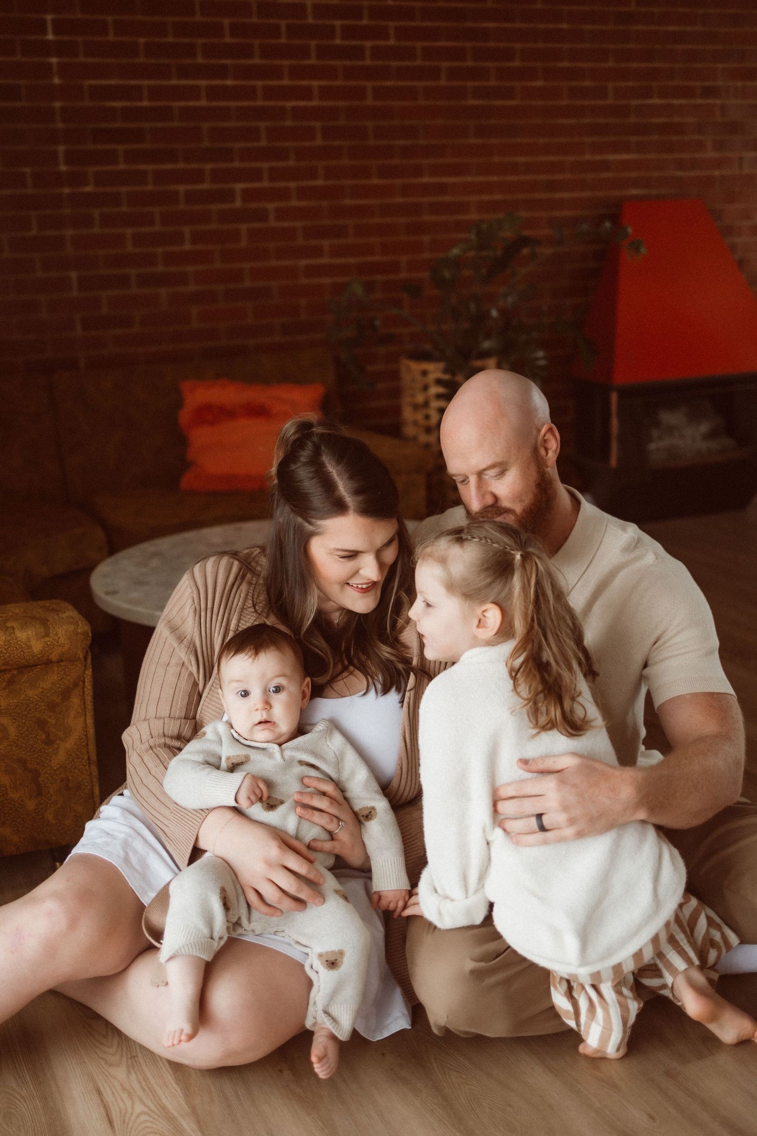 Family lounging together on floor against brick wall in neutral tones.