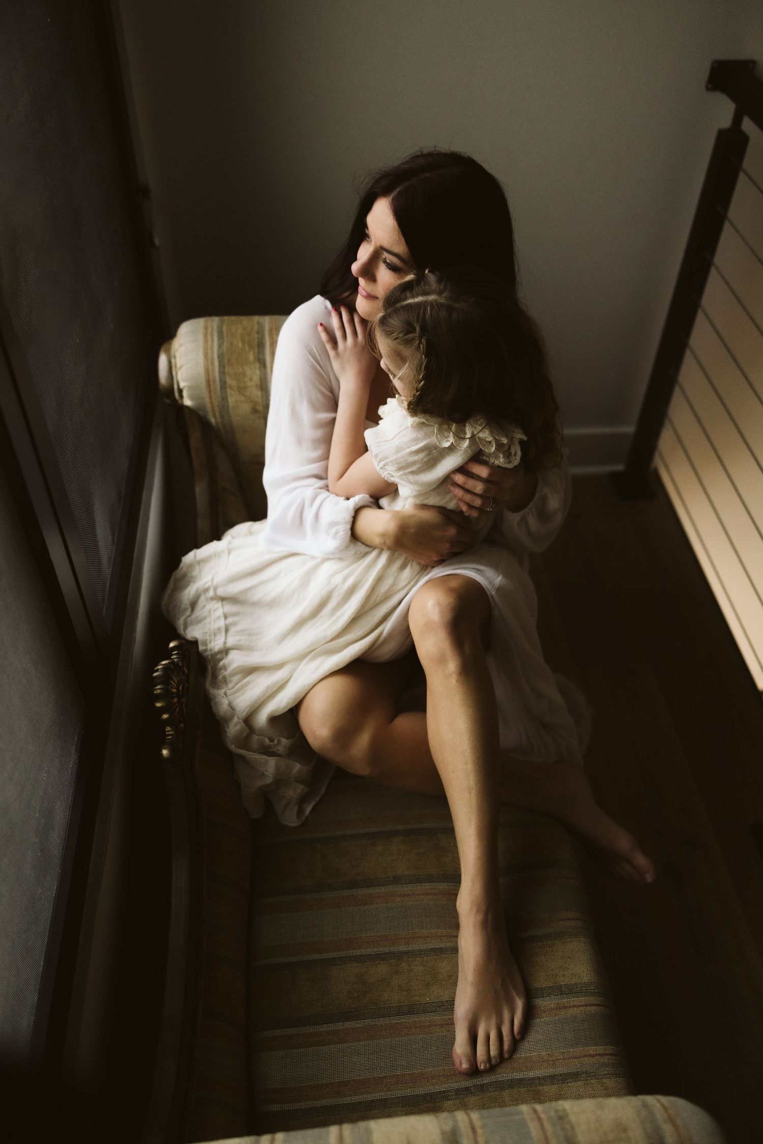 A tender embrace in white clothing while sitting on wooden stairs near a window.