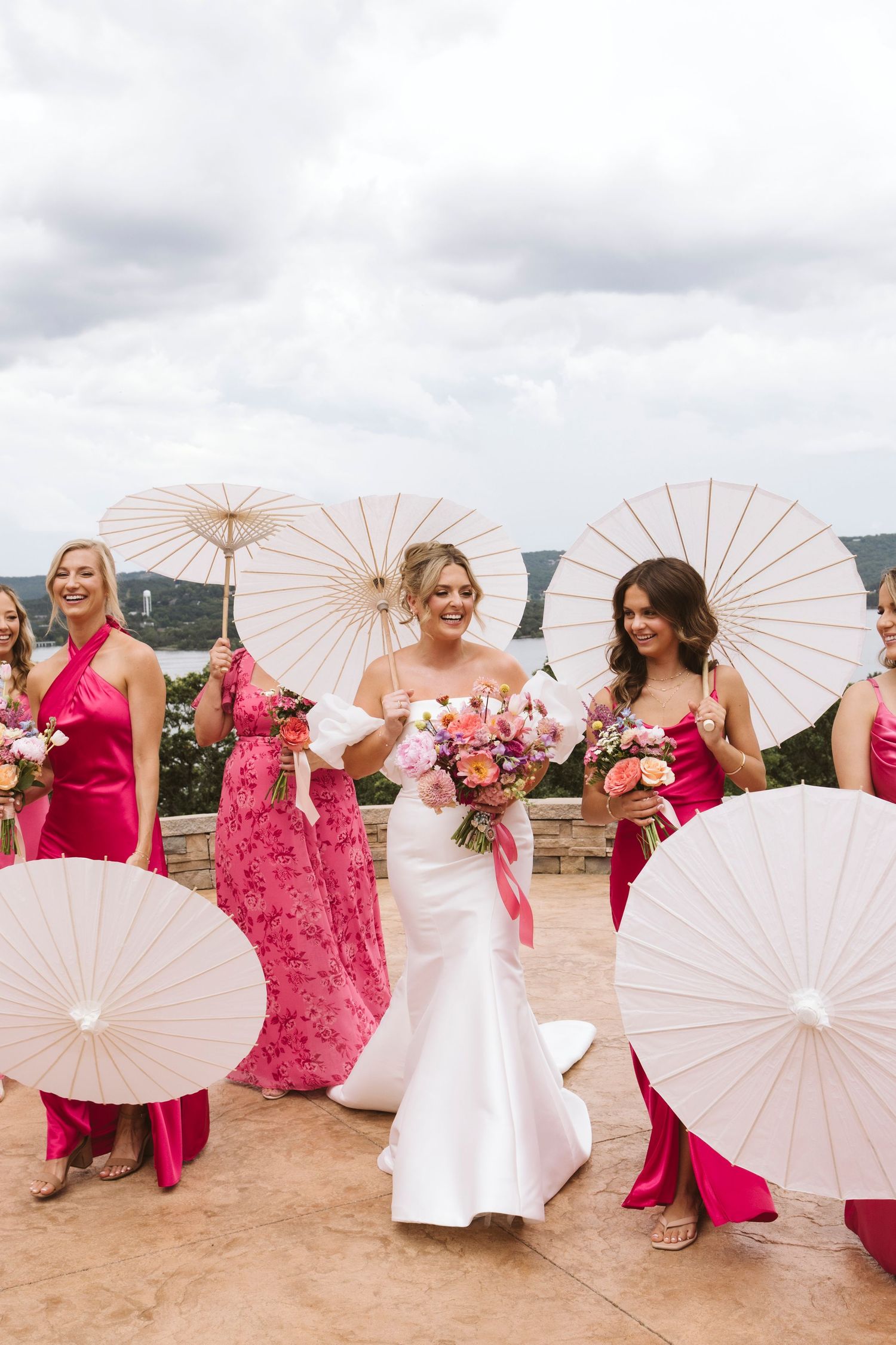 Wedding party with pink dresses and white parasols pose outdoors on a cloudy day.
