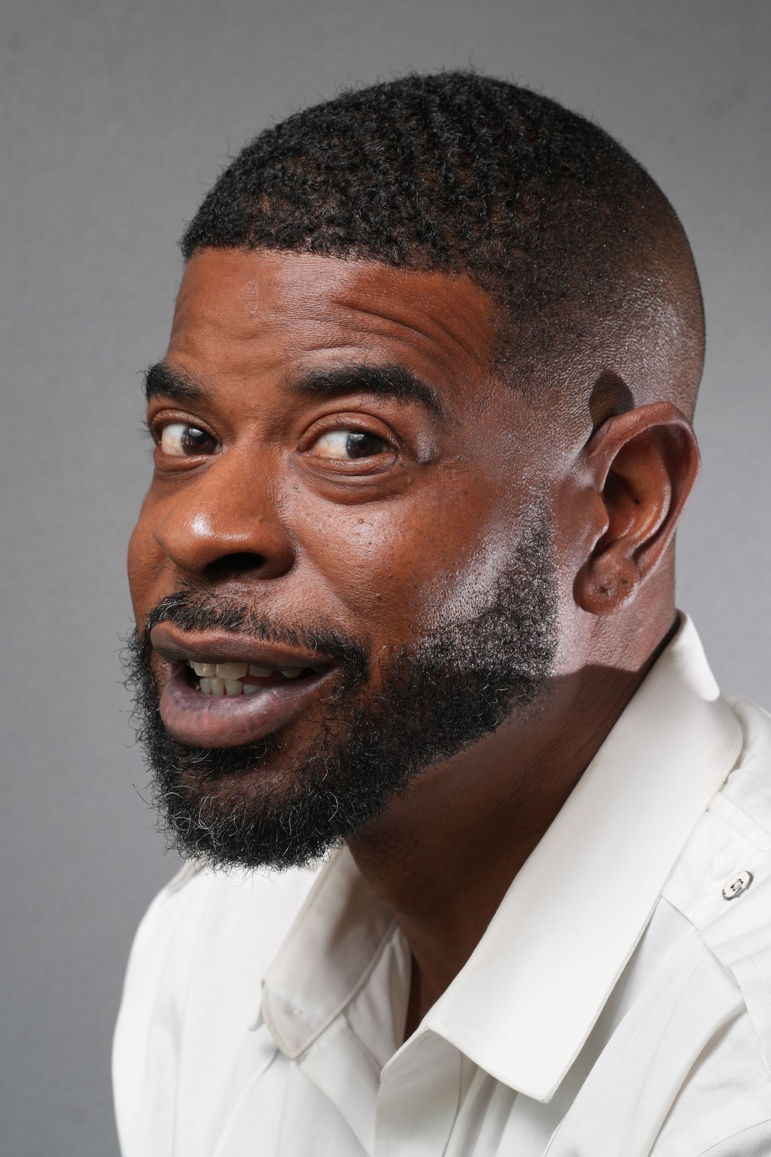 Cheerful person in white blazer with well-groomed beard makes playful expression against gray background.