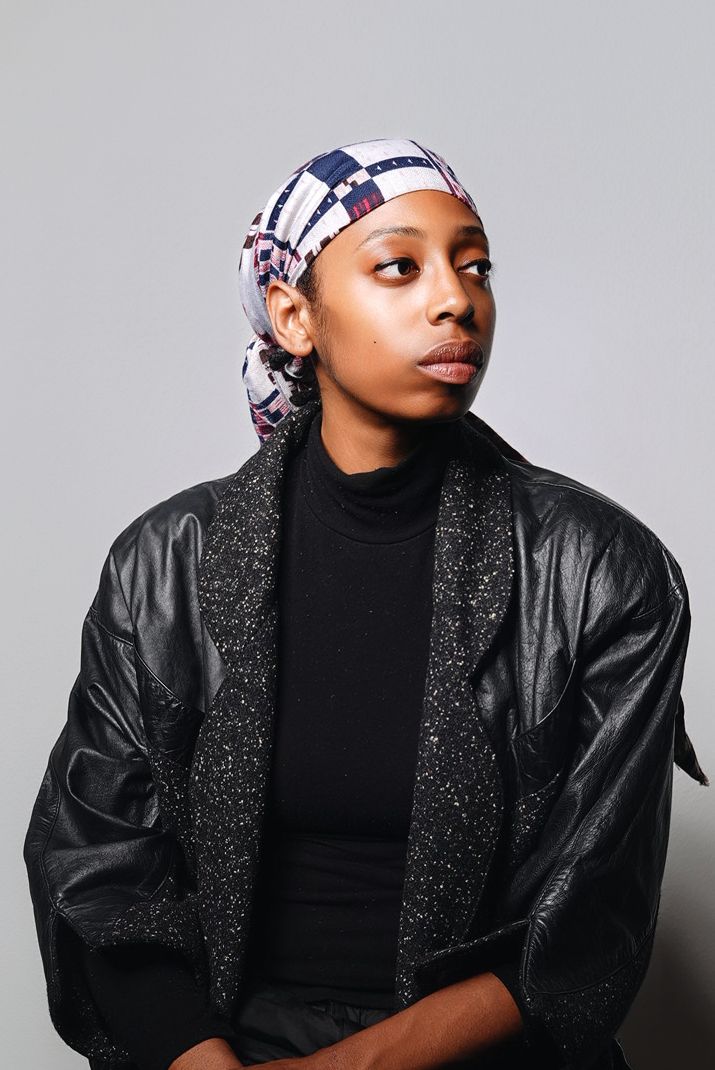 Artistic headshot of person wearing black turtleneck and patterned headwrap against gray background.