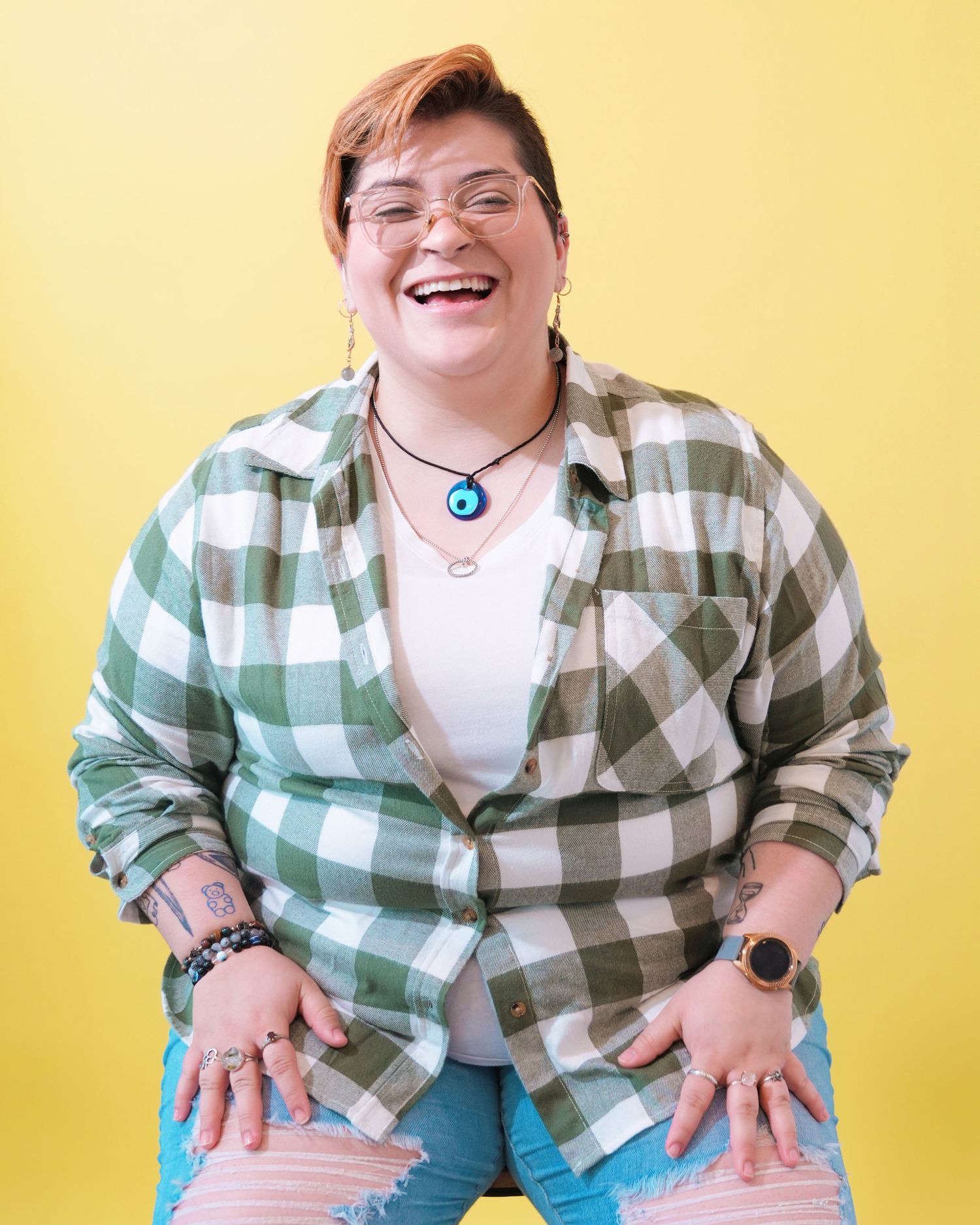Candid laughing pose in green plaid flannel and glasses against bright yellow wall.
