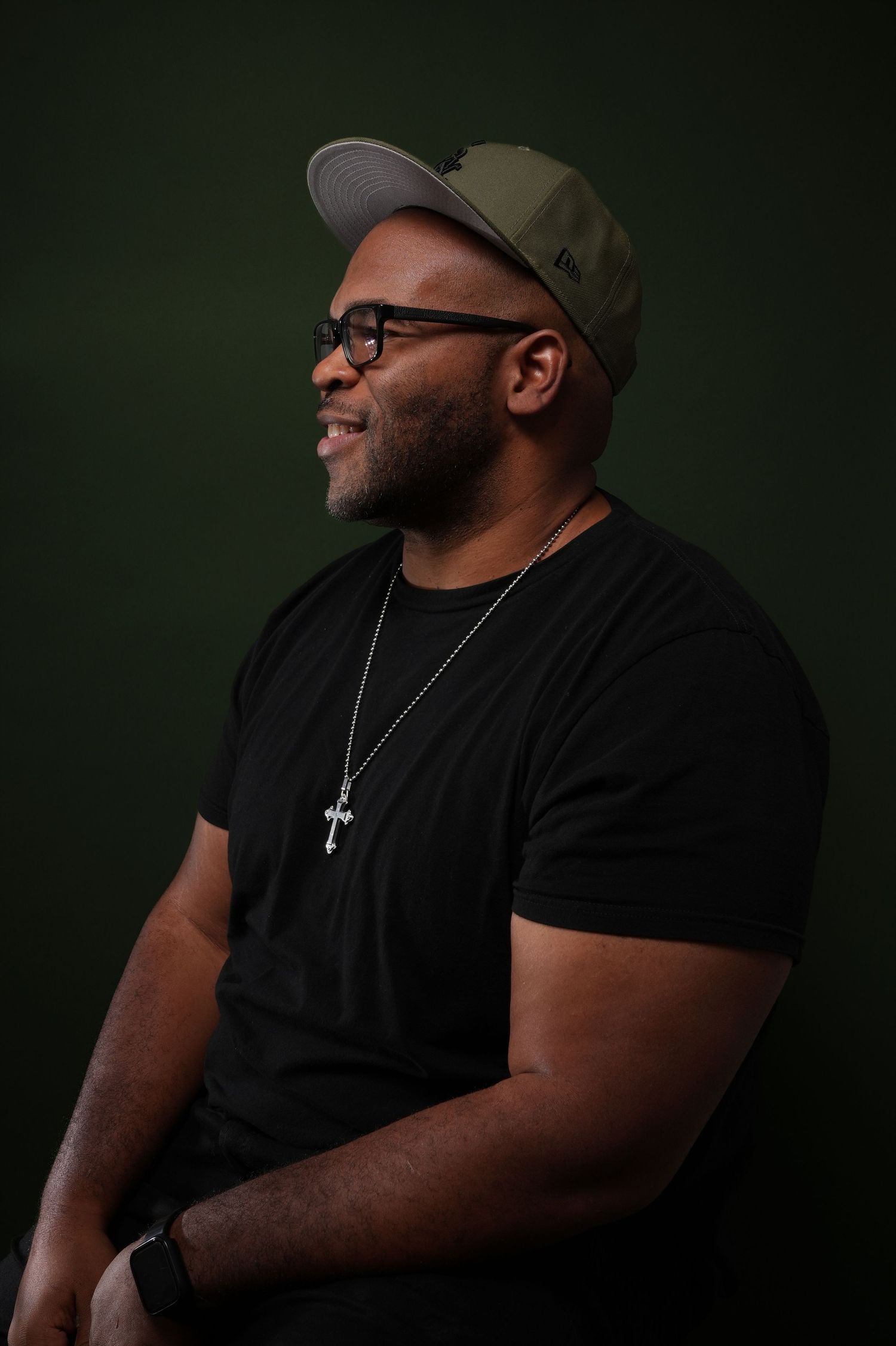 Studio portrait of a person in black t-shirt wearing baseball cap and cross necklace against dark background.