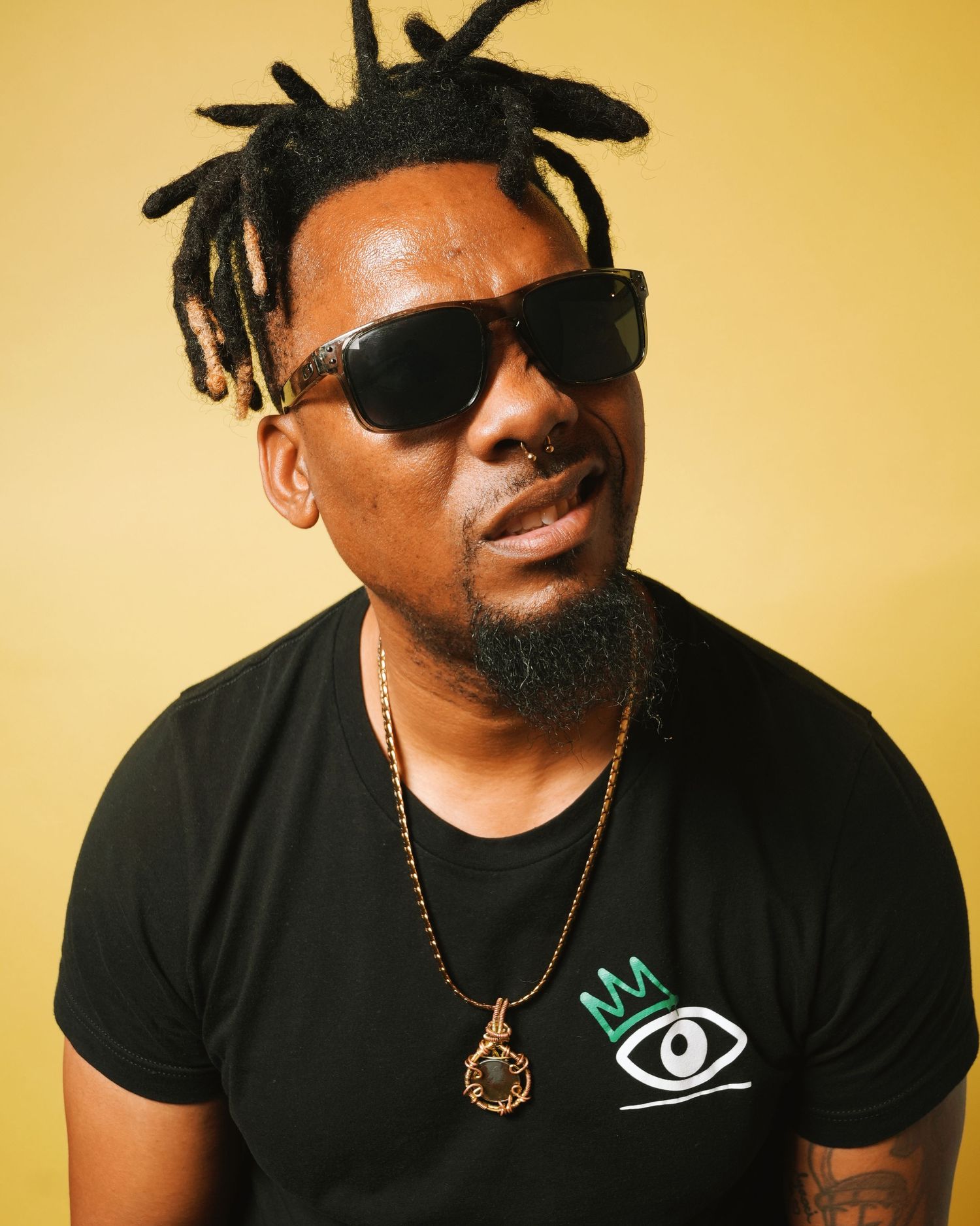 Studio portrait of someone in sunglasses and black t-shirt with locs hairstyle against yellow backdrop.