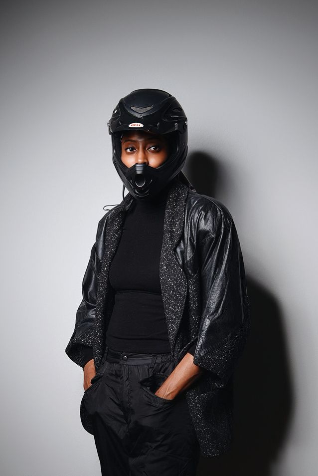 Studio portrait of someone wearing a black motorcycle helmet and leather jacket against a grey background.