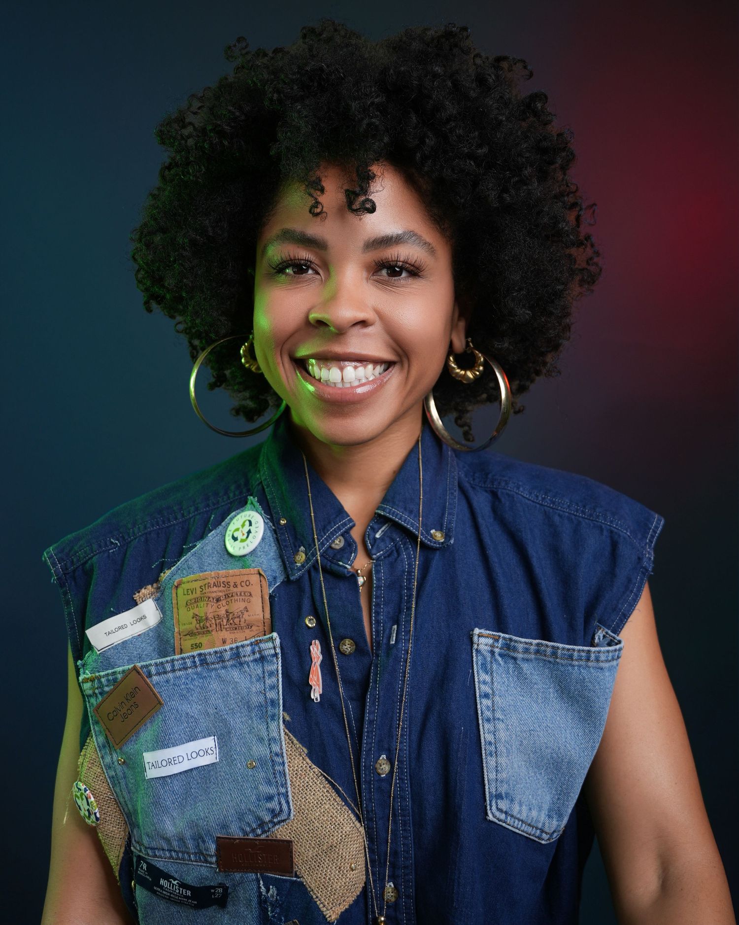 Smiling young person in denim sleeveless jacket against colorful background wearing gold hoop earrings and natural curls.