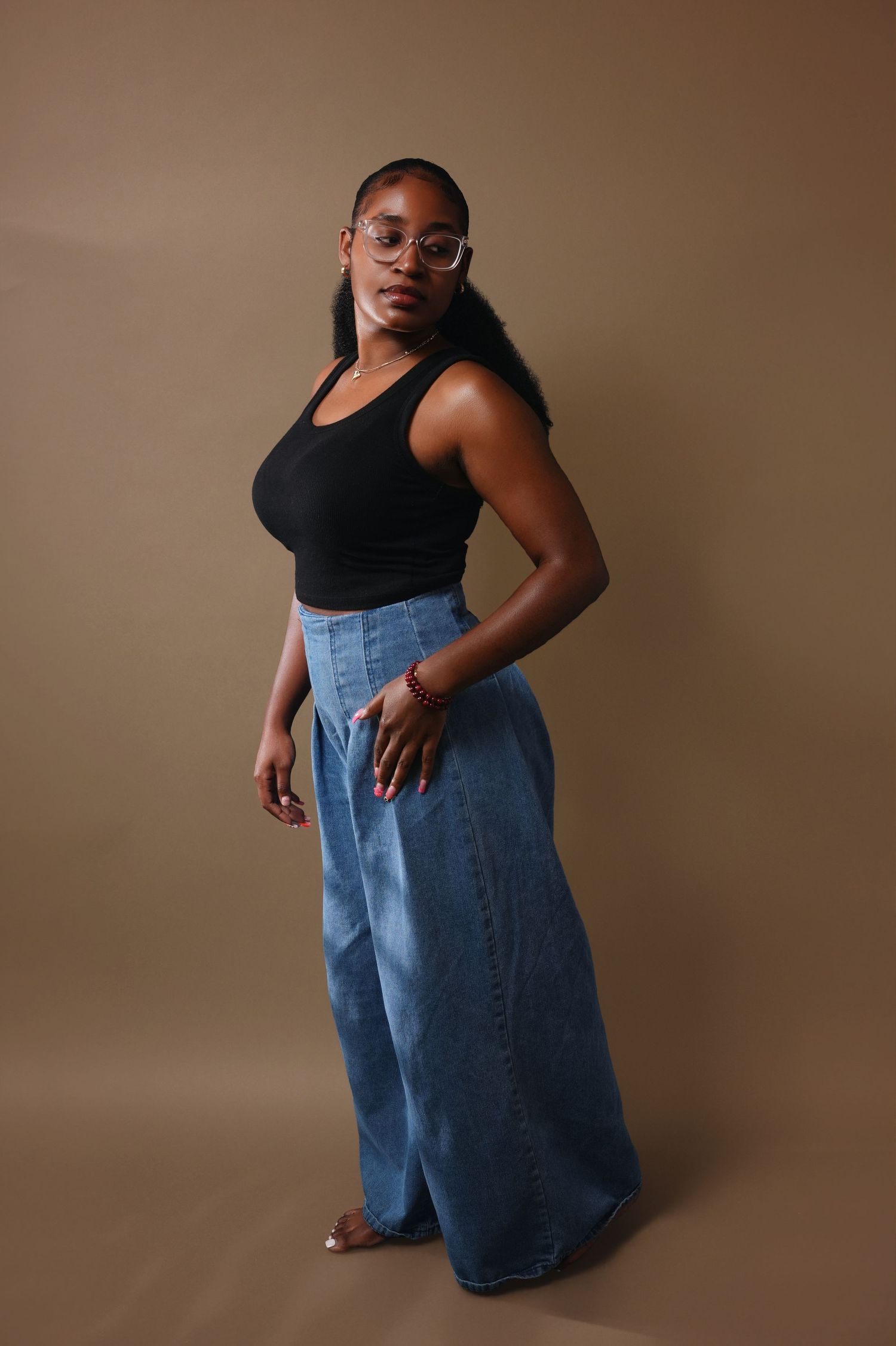 Full body portrait of a person in black tank top and wide-leg jeans against brown studio backdrop.